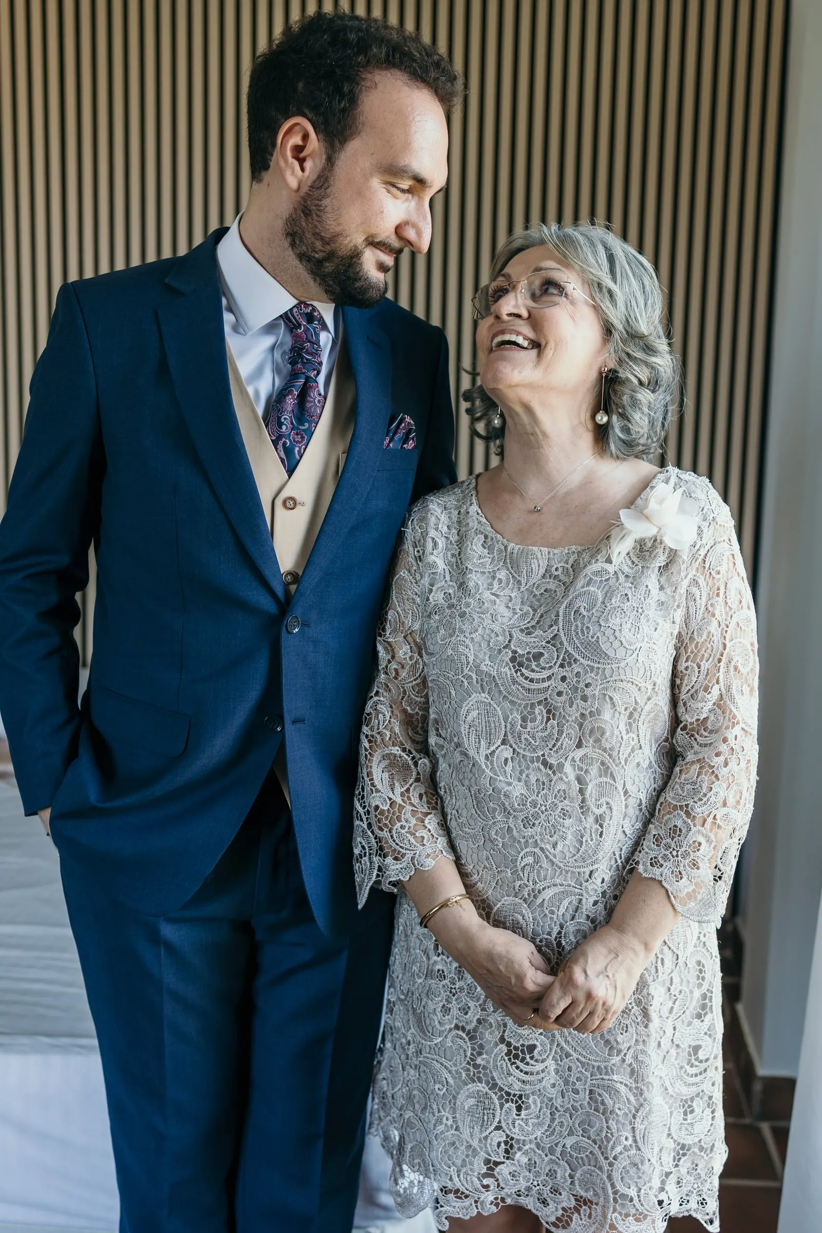 Groom in a navy suit and a woman in a grey lace dress smiling at each other before the wedding