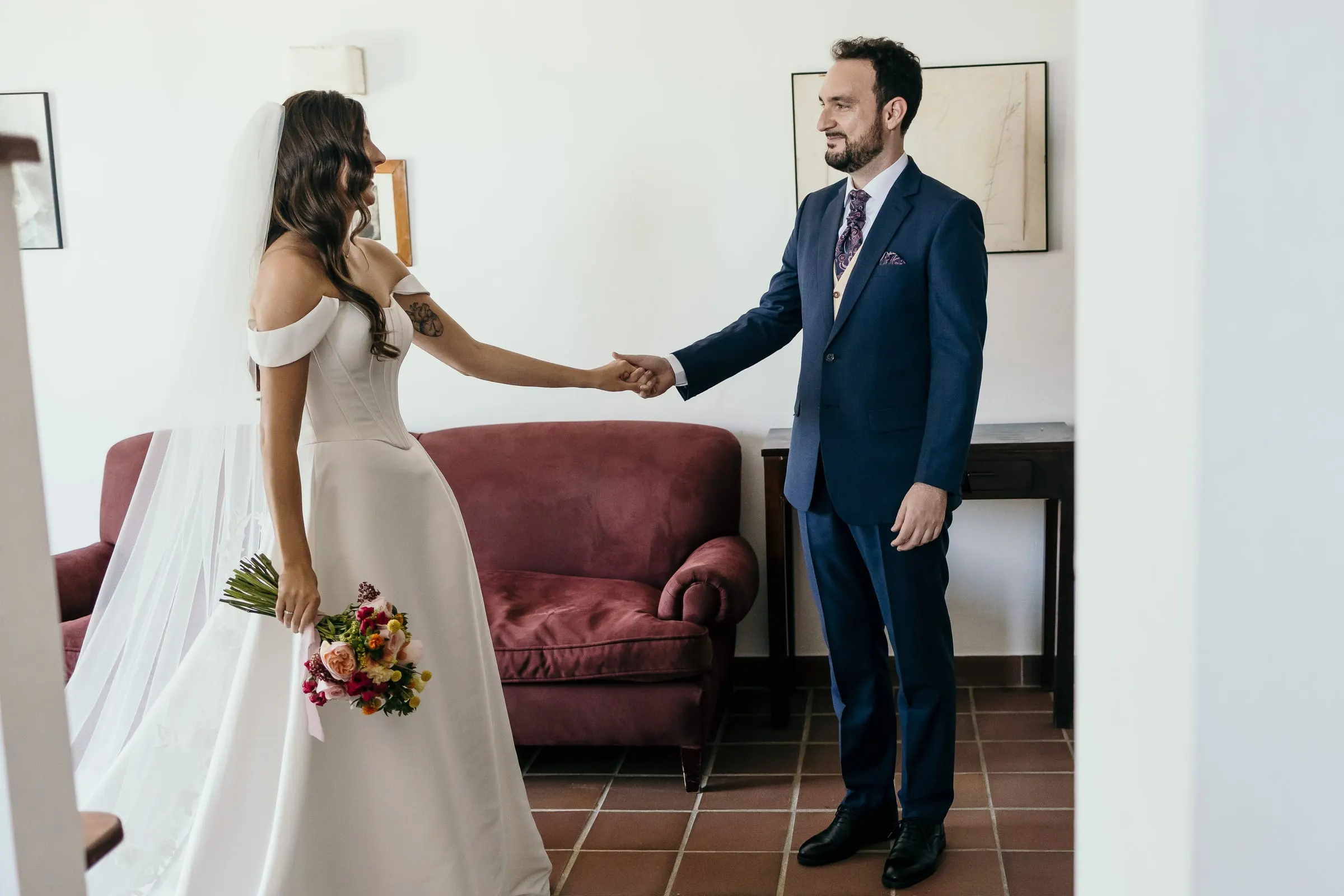Bride and groom reaching for each other's hand at their first look, a red sofa behind