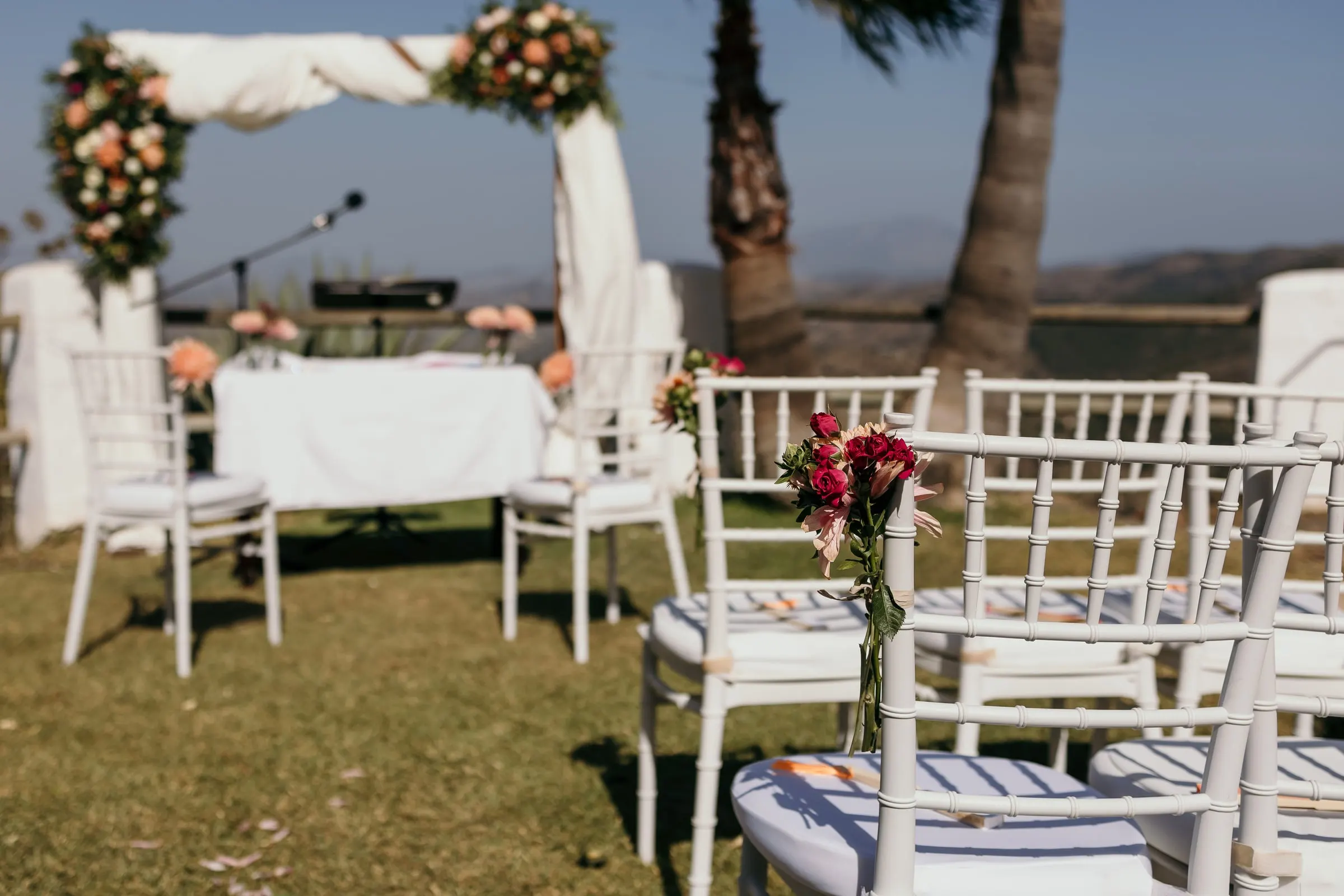 Ceremony chairs tied with pink flowers, with the floral arch and palm trees behind