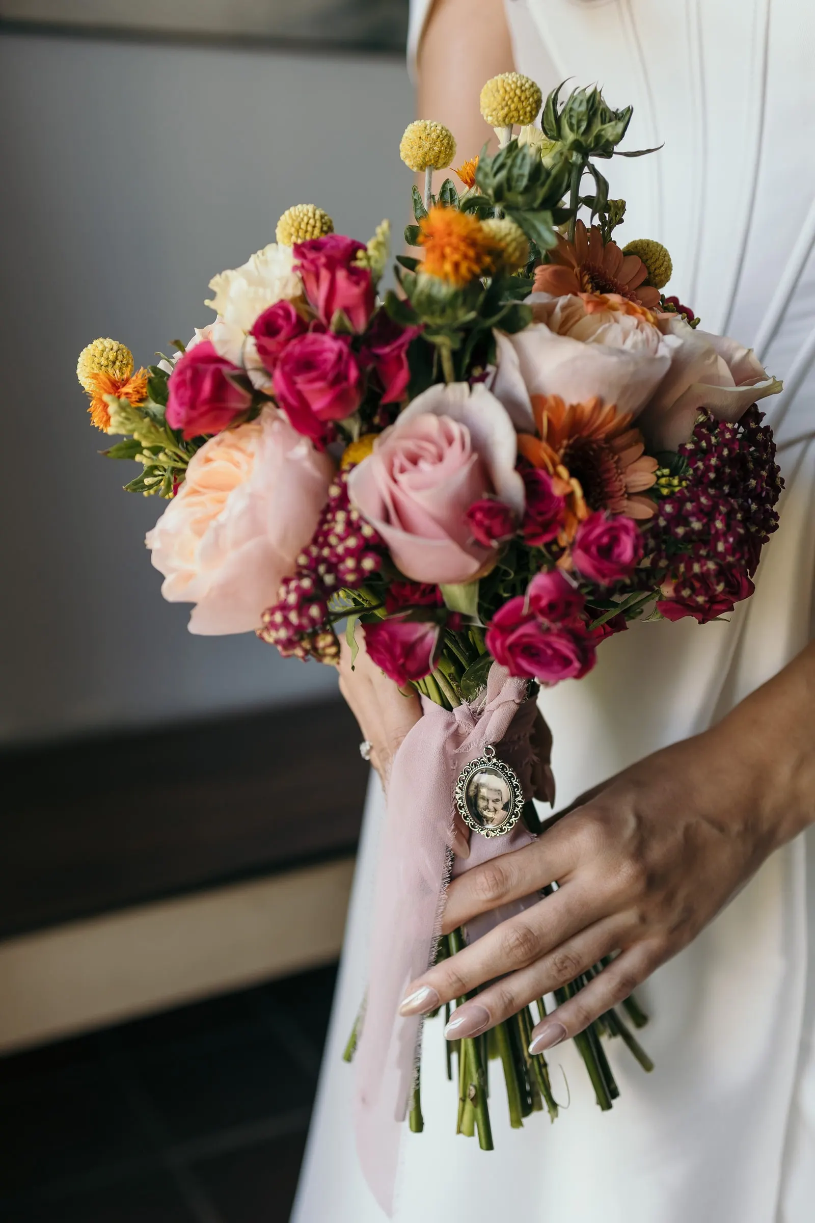 Close-up of the bridal bouquet of roses, craspedia and orange blooms with a pink ribbon and a small charm