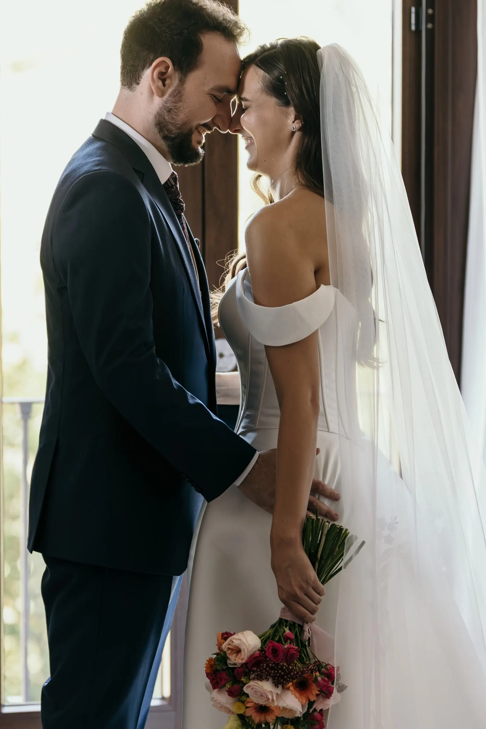 Bride and groom forehead to forehead by a sunlit window, bouquet in hand