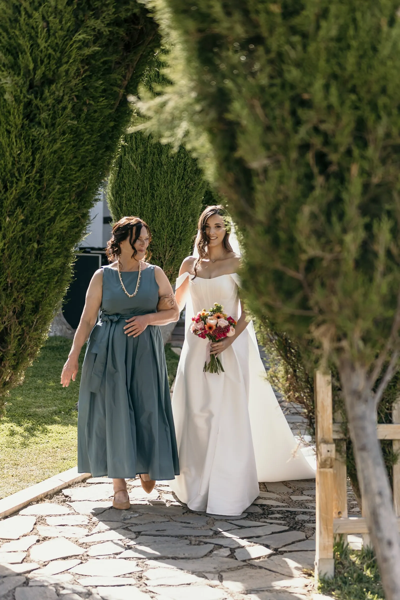 Bride walking arm in arm with the woman in teal between cypress trees towards the ceremony