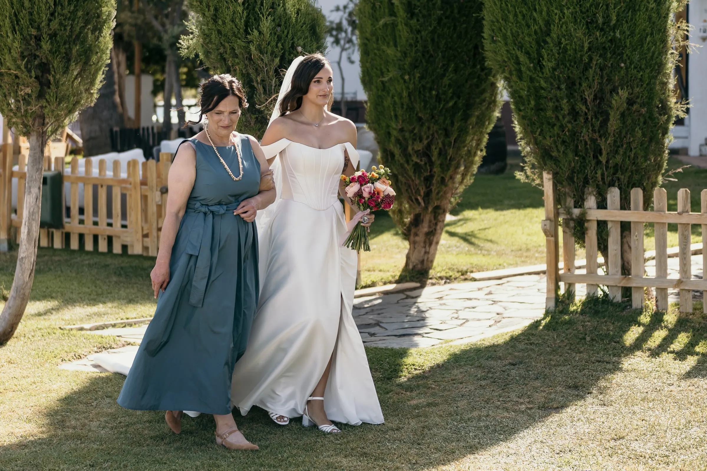 Bride and the woman in teal crossing the garden past a wooden fence towards the aisle