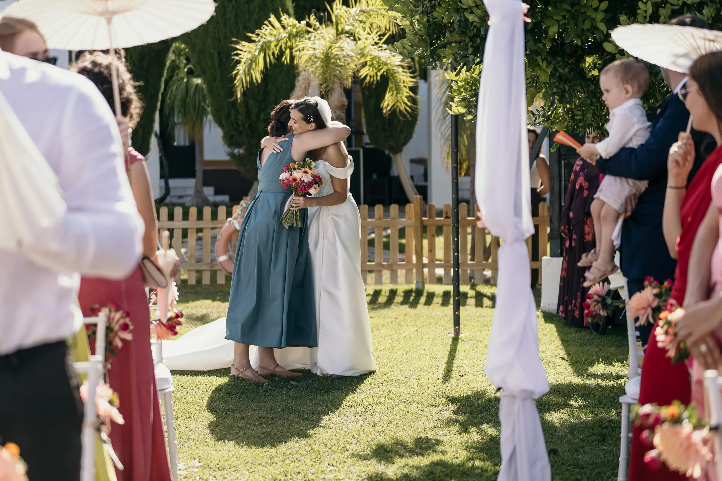 Bride and the woman in teal hugging at the end of the aisle as guests with paper parasols look on