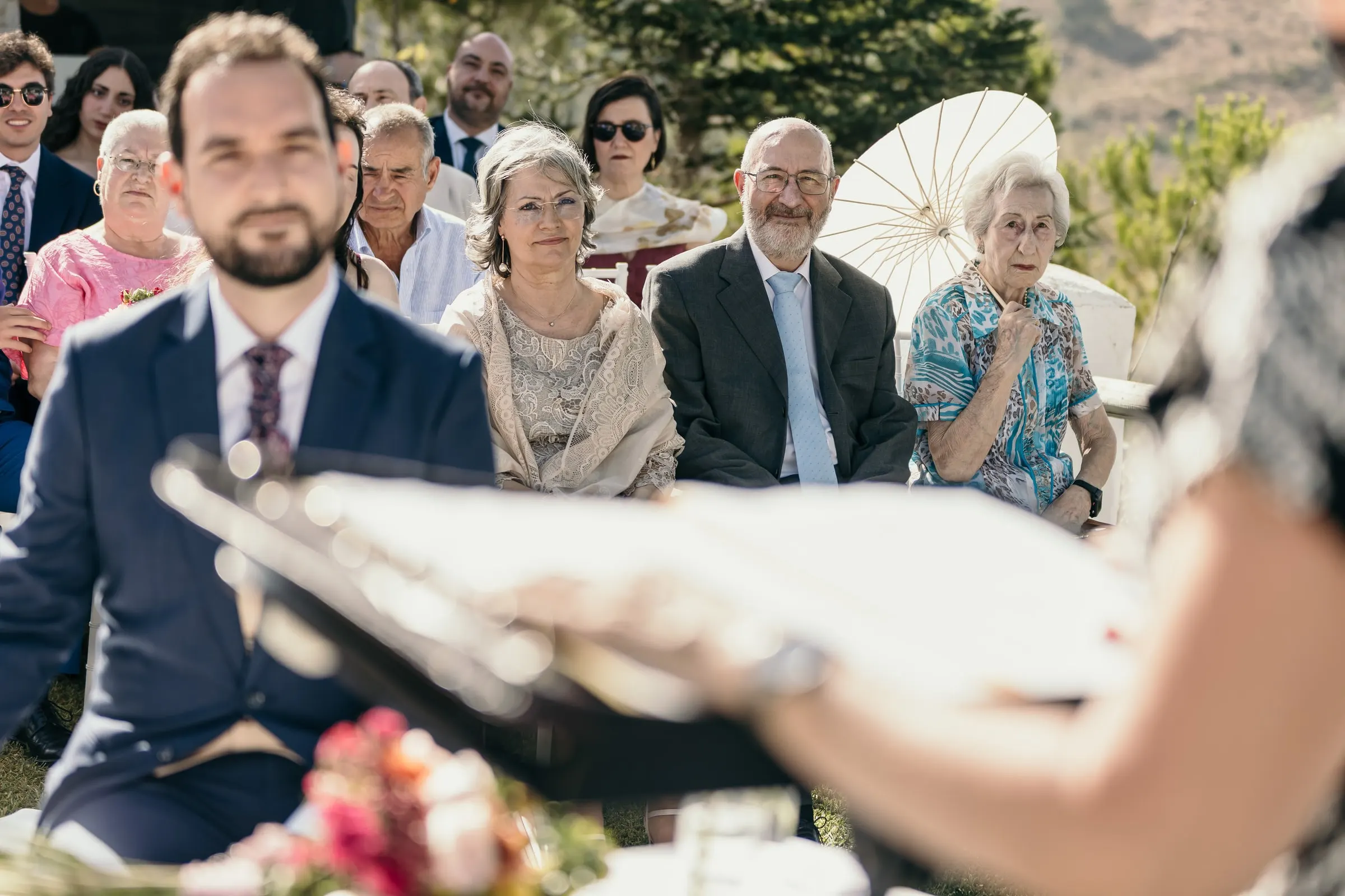 Groom listening to a reading, older guests and a paper parasol behind him