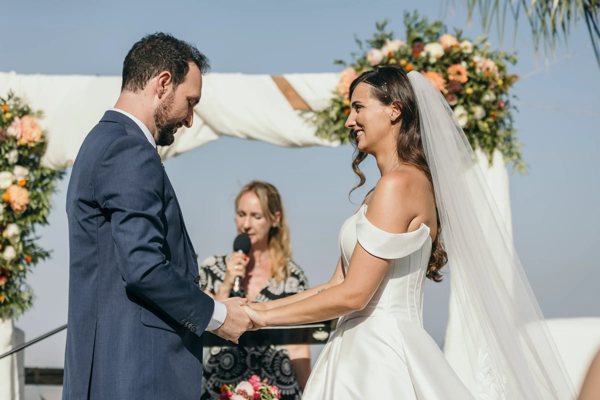 Bride and groom holding hands under the floral arch as the celebrant speaks