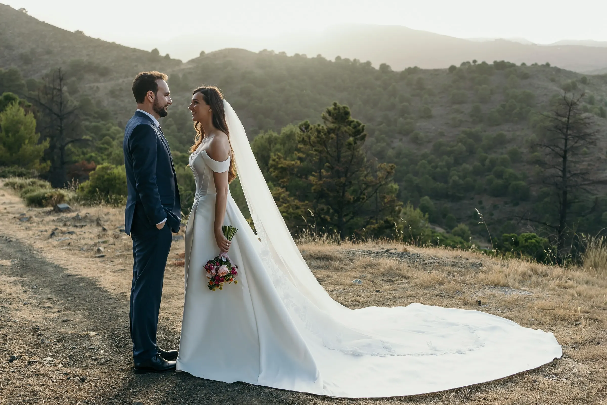 Los novios frente a frente en un camino en lo alto de la sierra con capas de montañas detrás, boda en la Costa del Sol