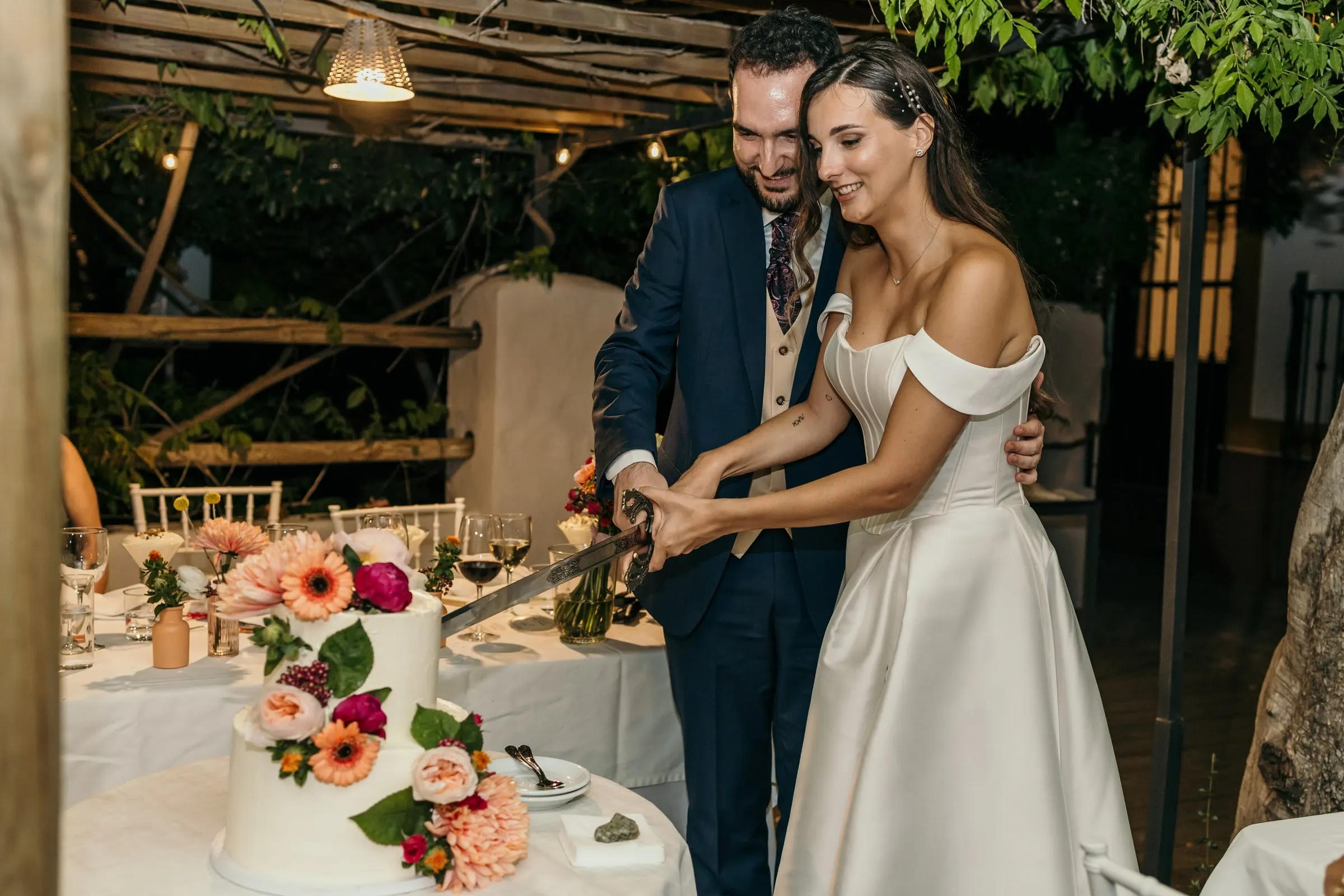 Bride and groom cutting their flower-topped cake with a sword under a vine pergola