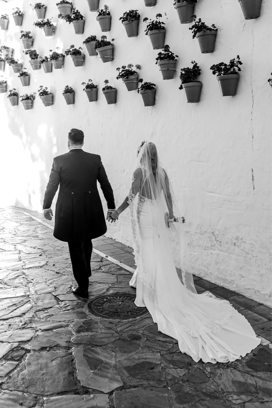 Los novios suben de la mano por una calle encalada llena de macetas, en blanco y negro, Costa del Sol