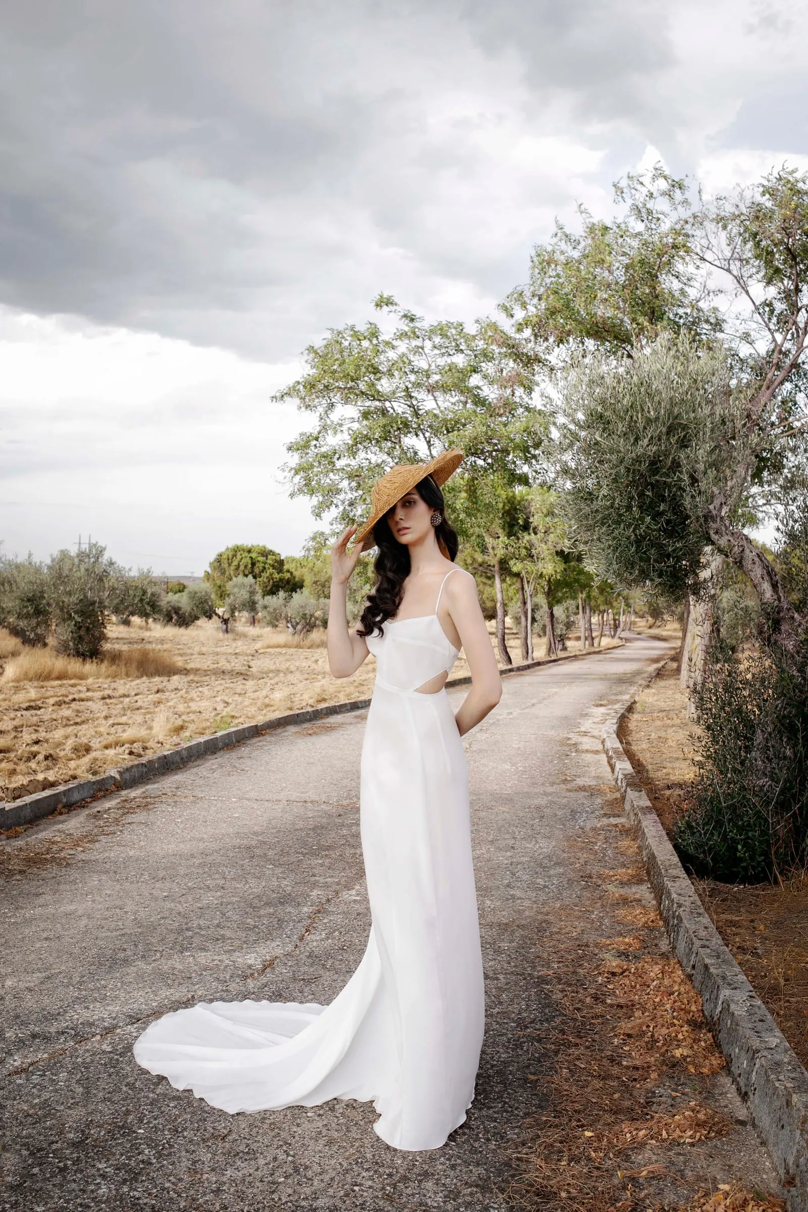 Bride in a wide straw hat and long train standing on a tree-lined country lane, Costa del Sol