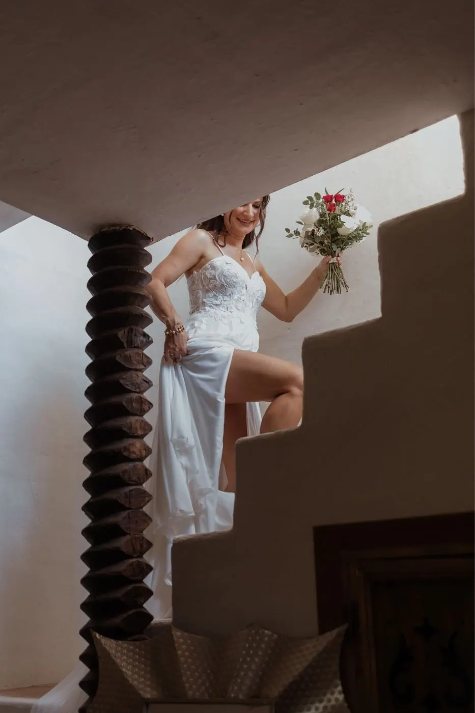 La novia se recoge la falda al subir una escalera blanca, ramo en mano, vista desde abajo, Costa del Sol