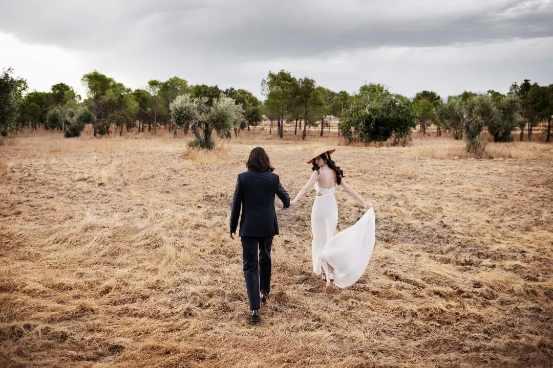Bride in a straw hat and groom walk hand in hand across a golden field towards olive trees, Costa del Sol