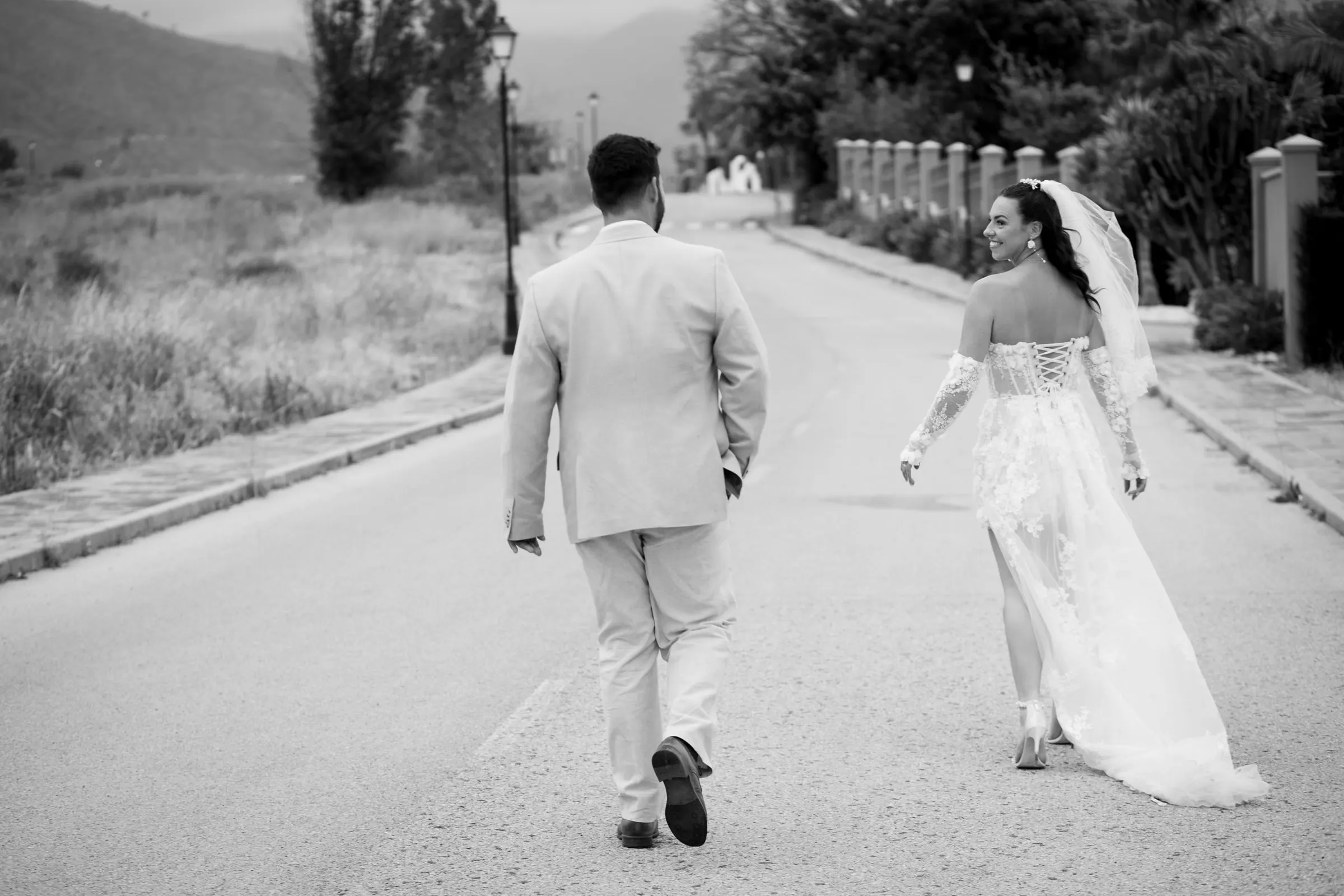 Blanco y negro de los novios caminando por el camino de Hacienda San José, la novia mirando atrás con una sonrisa, Mijas