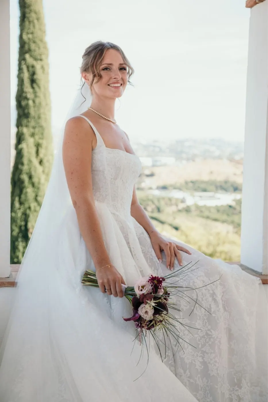 La novia, sentada en un alféizar blanco con su ramo, sonríe con el valle detrás, Hacienda San José, Mijas