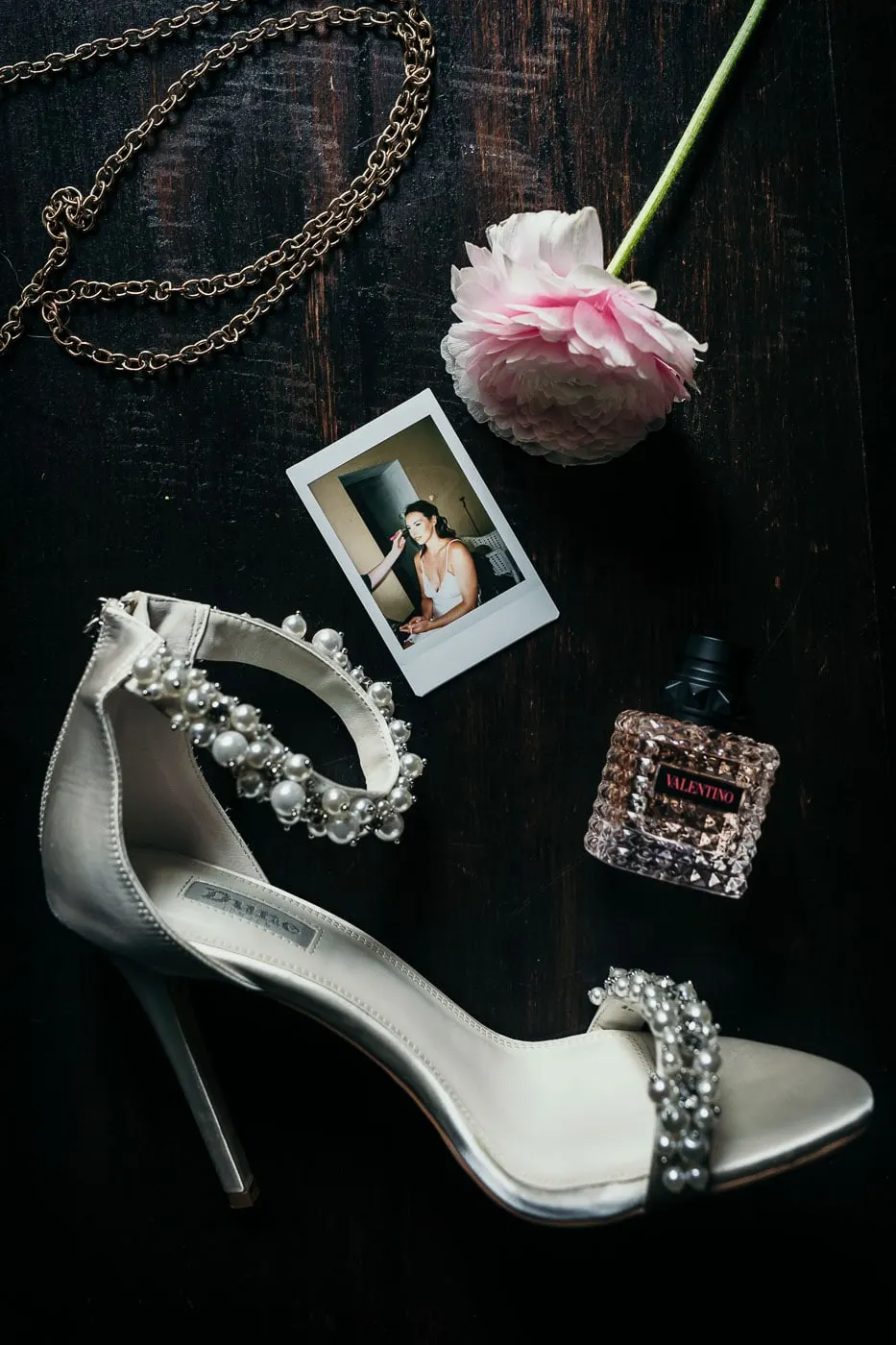 Tacones con perlas, un ranúnculo rosa, perfume y una Polaroid sobre madera oscura, Hacienda San José, Mijas