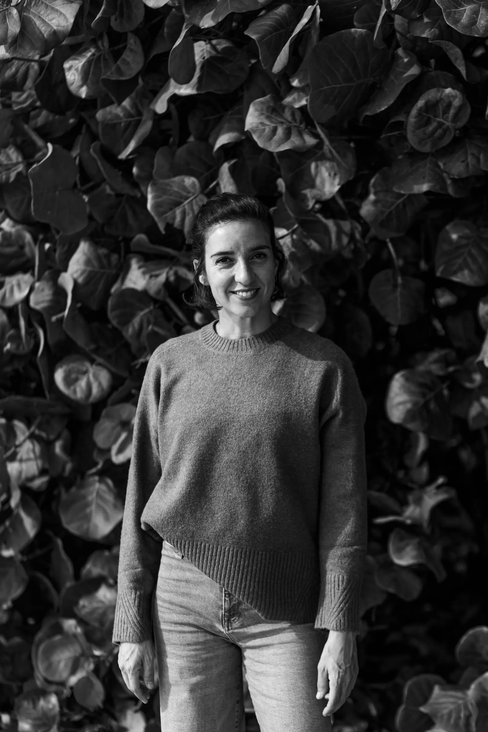 Retrato en blanco y negro de Tamara, videógrafa de Picart, sonriendo ante un muro de hojas, Estepona
