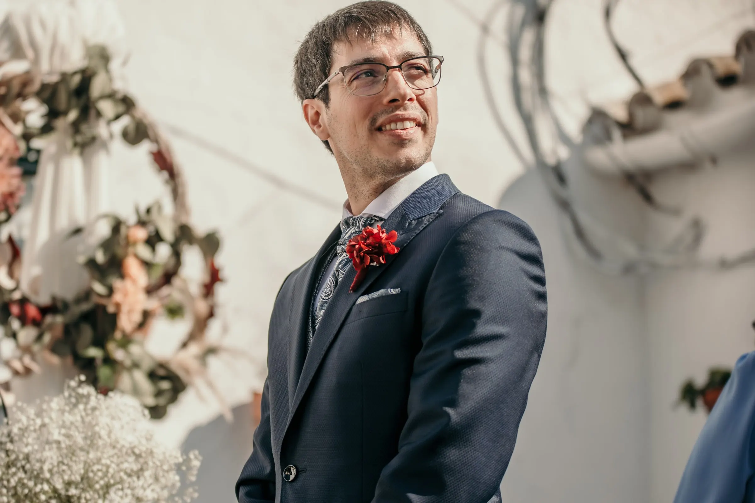 Groom in a navy suit with a red buttonhole smiling as he waits beside a whitewashed wall