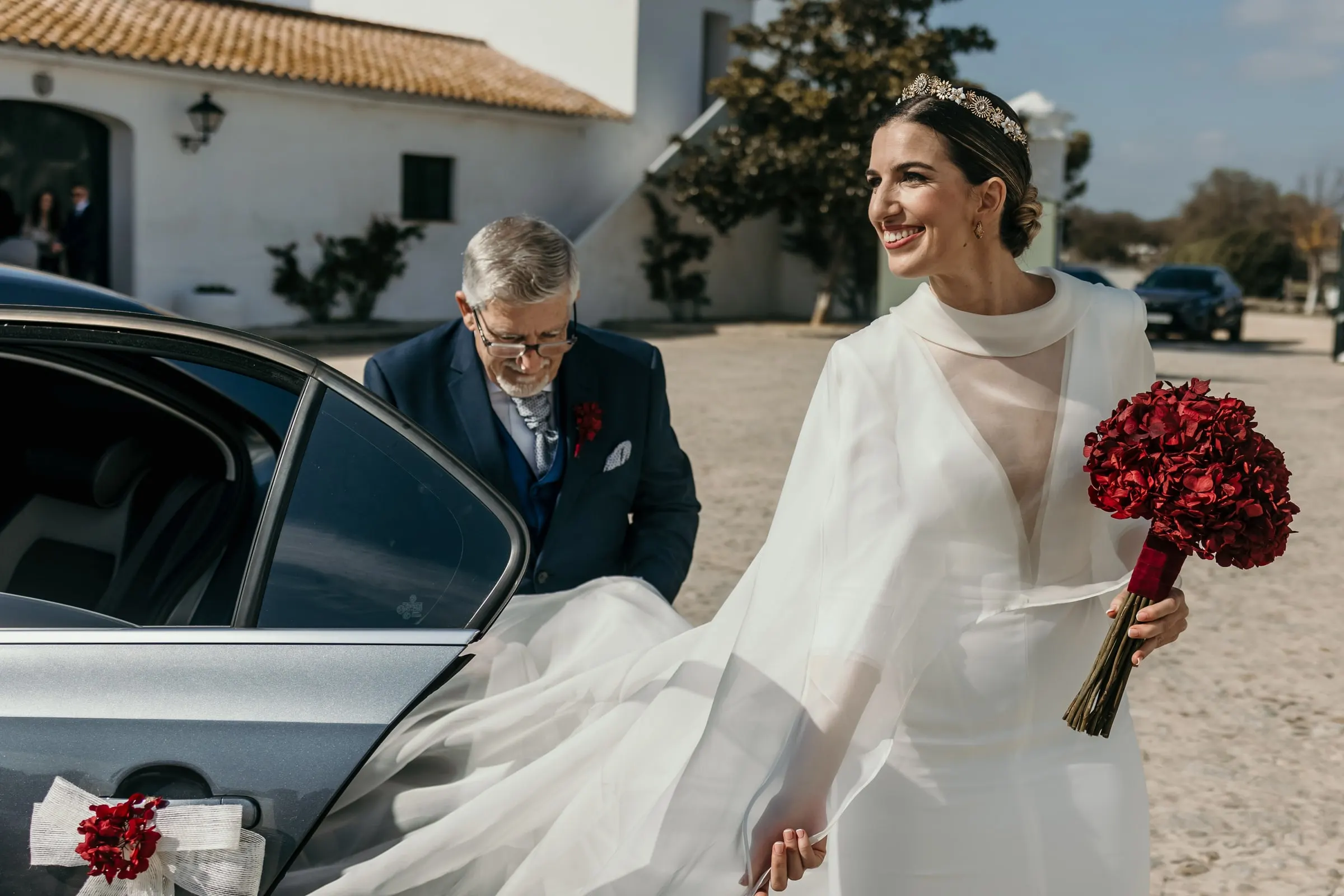 Bride stepping out of the car with her red rose bouquet and long veil in front of the white cortijo buildings