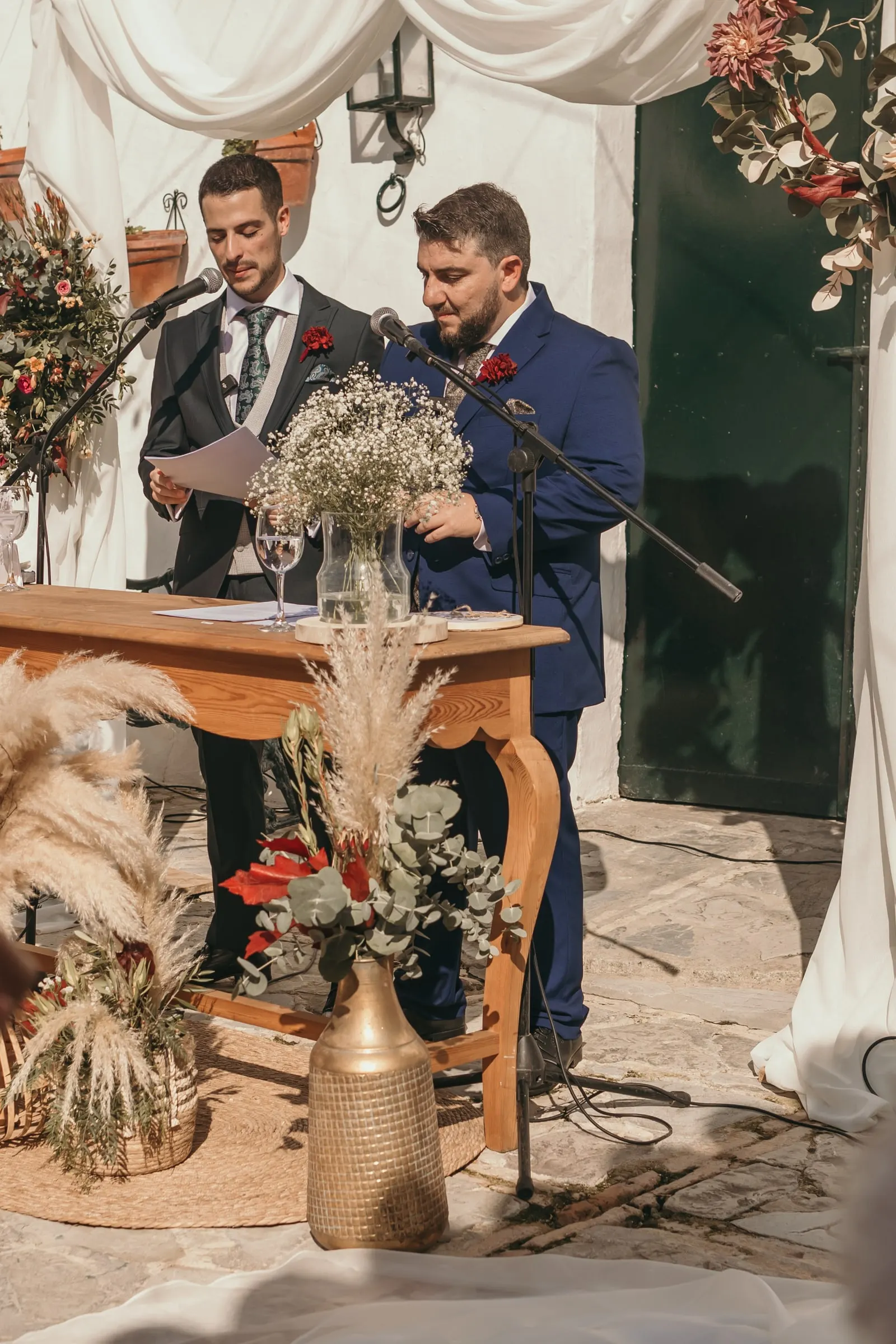 Two friends reading at a microphone behind a wooden table with gypsophila and pampas grass, cortijo ceremony