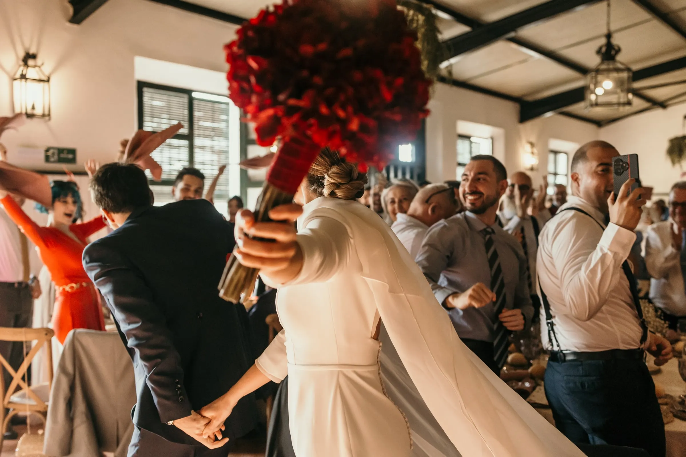 Bride raising her red rose bouquet as guests film the couple's entrance to dinner