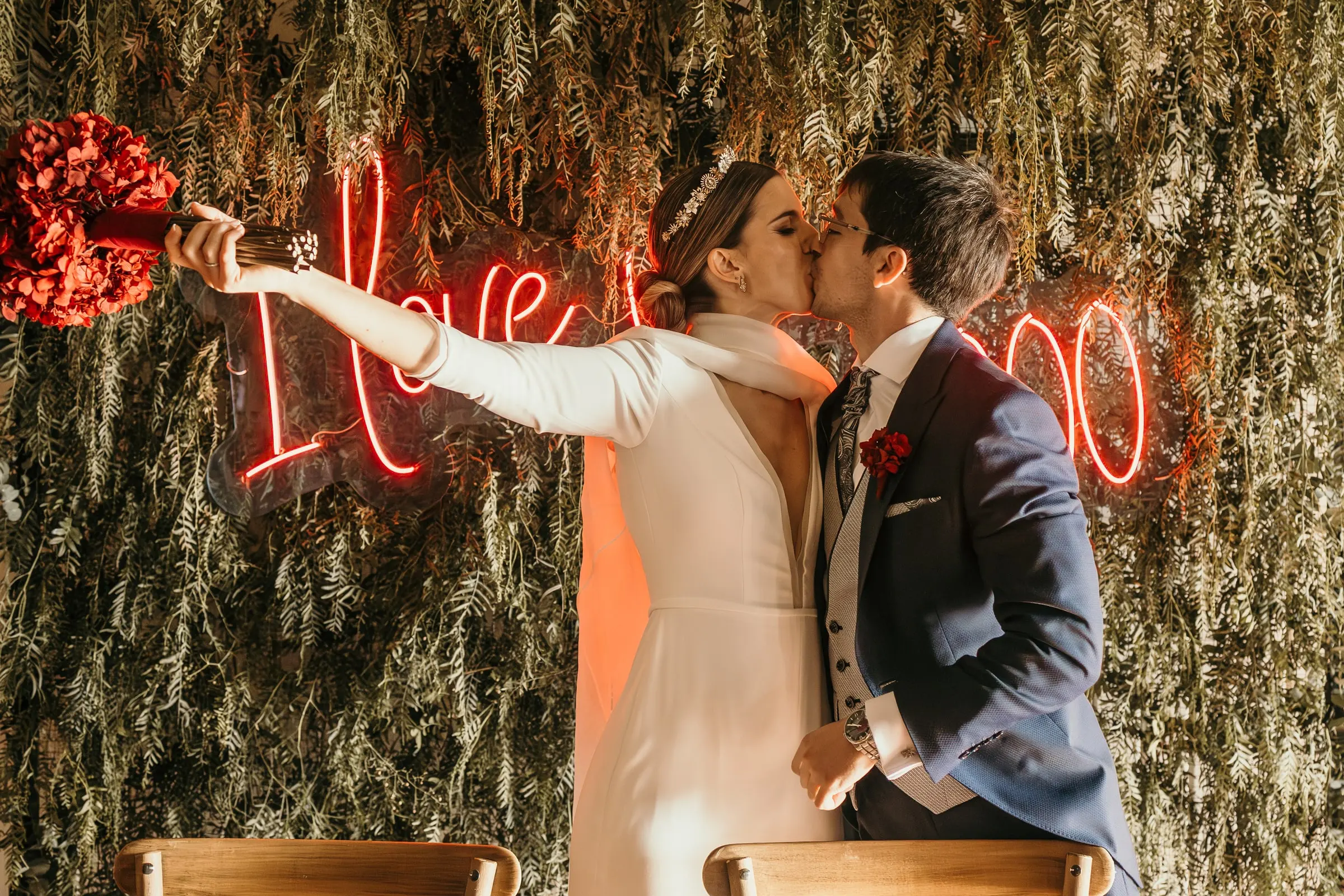 Bride and groom kissing in front of a pink neon sign on a green hedge wall
