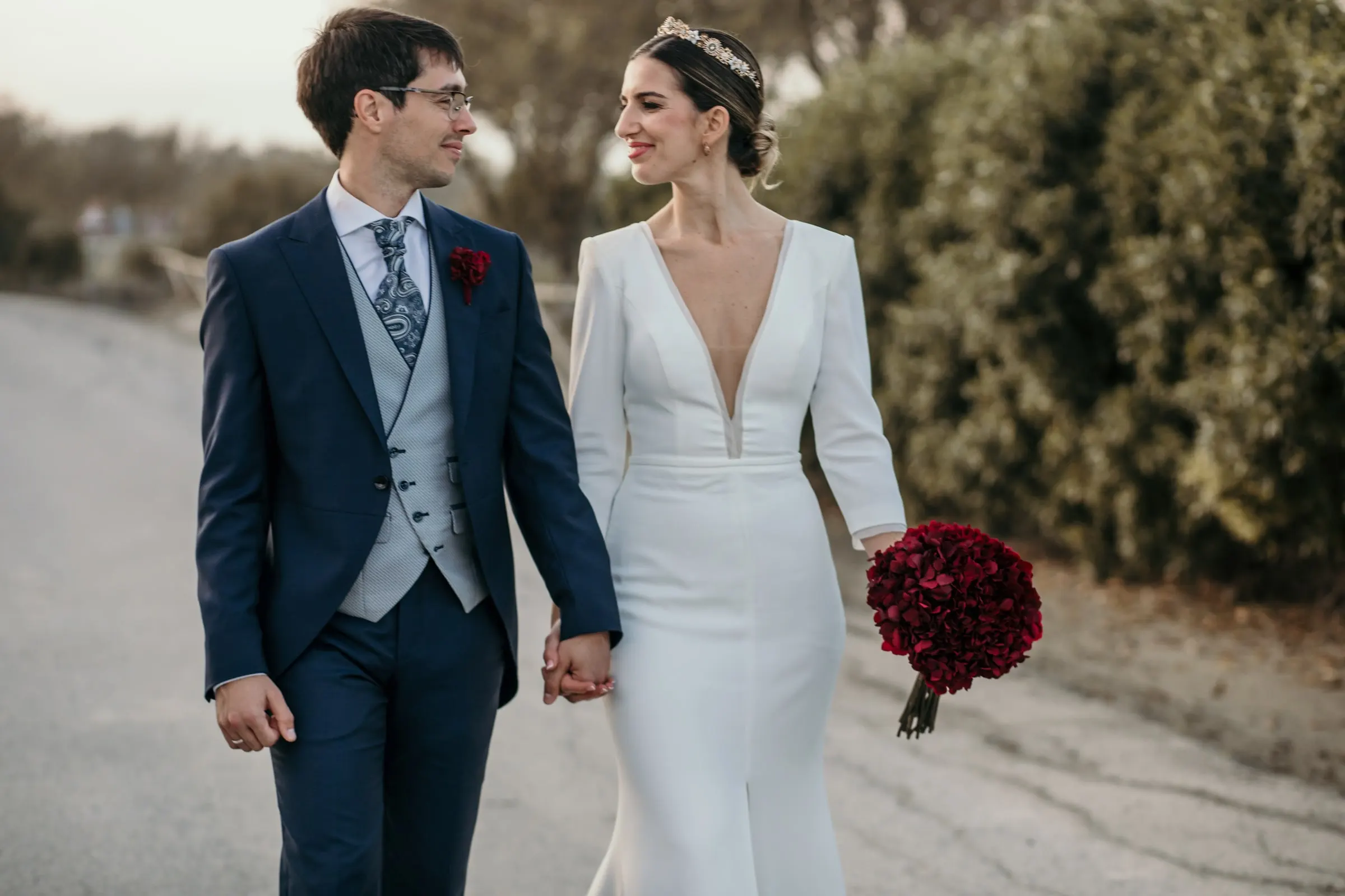 Bride and groom walking hand in hand along the road with her red bouquet at her side