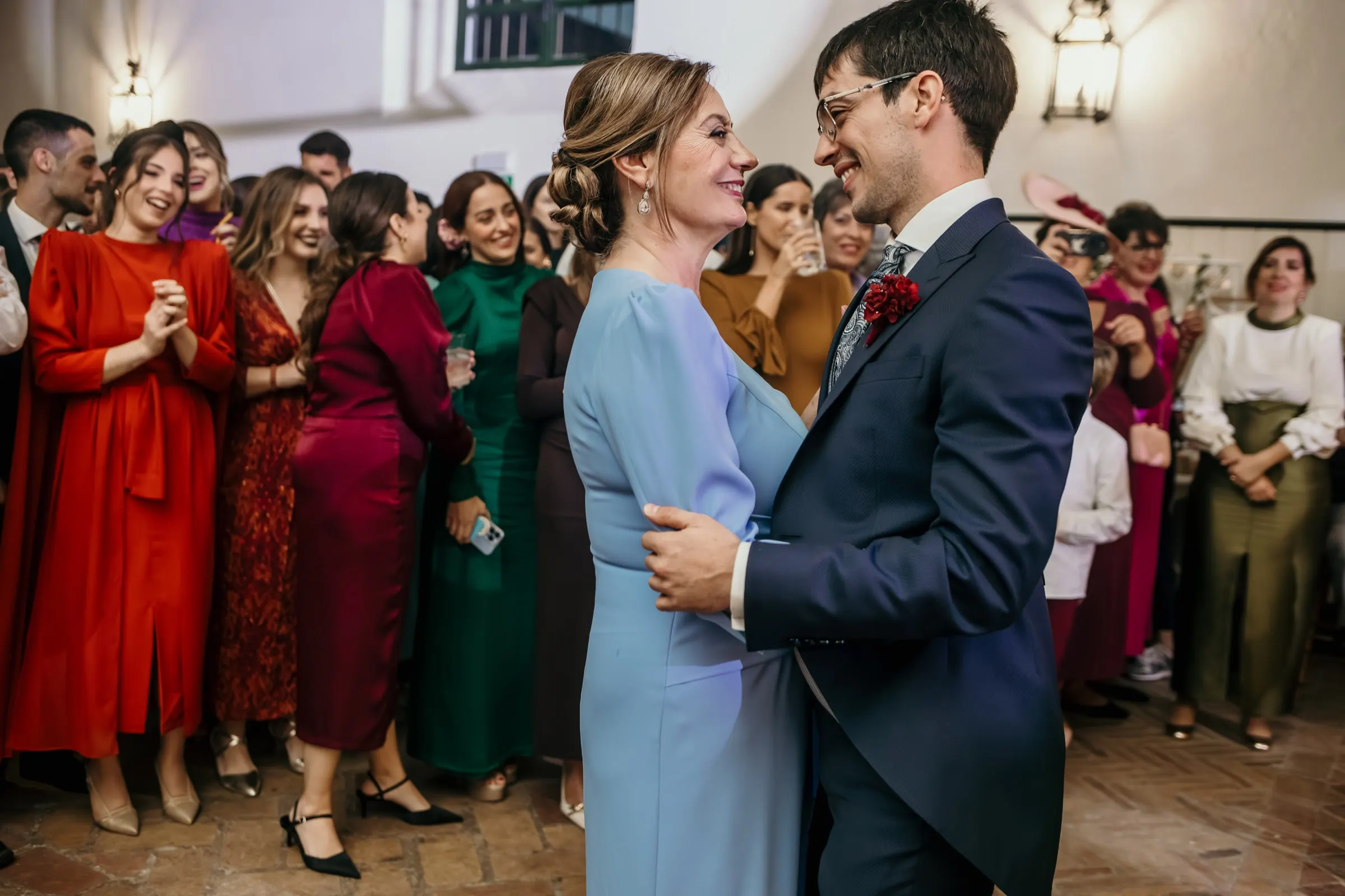 Groom dancing with his mother in the banquet hall, surrounded by guests in colourful dresses