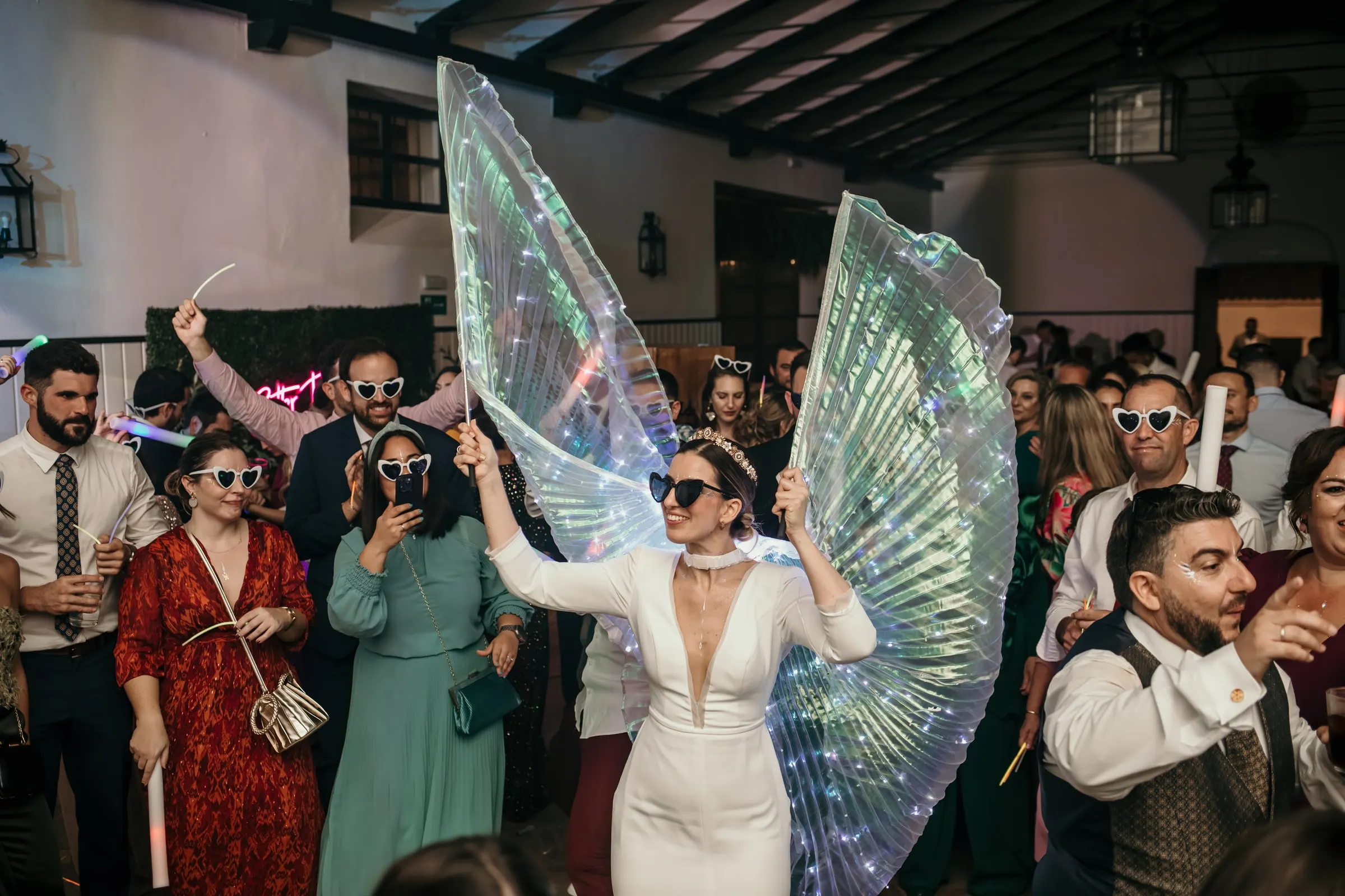 Bride with LED wings in the middle of the dance floor surrounded by guests in sunglasses