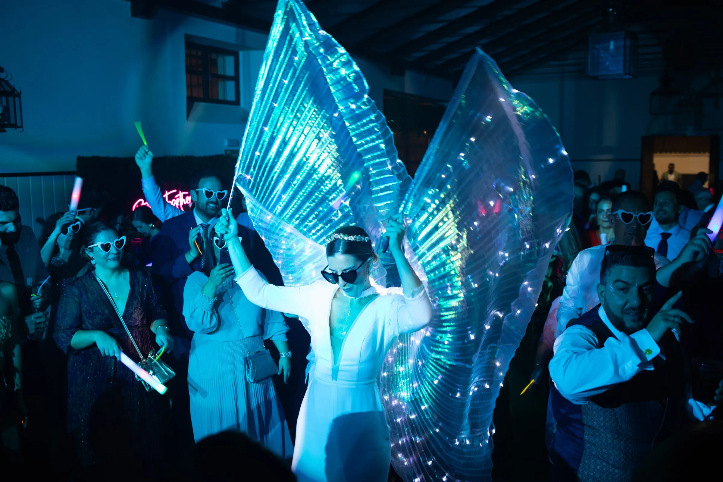 Bride's LED wings glowing under blue light in the crowded wedding party