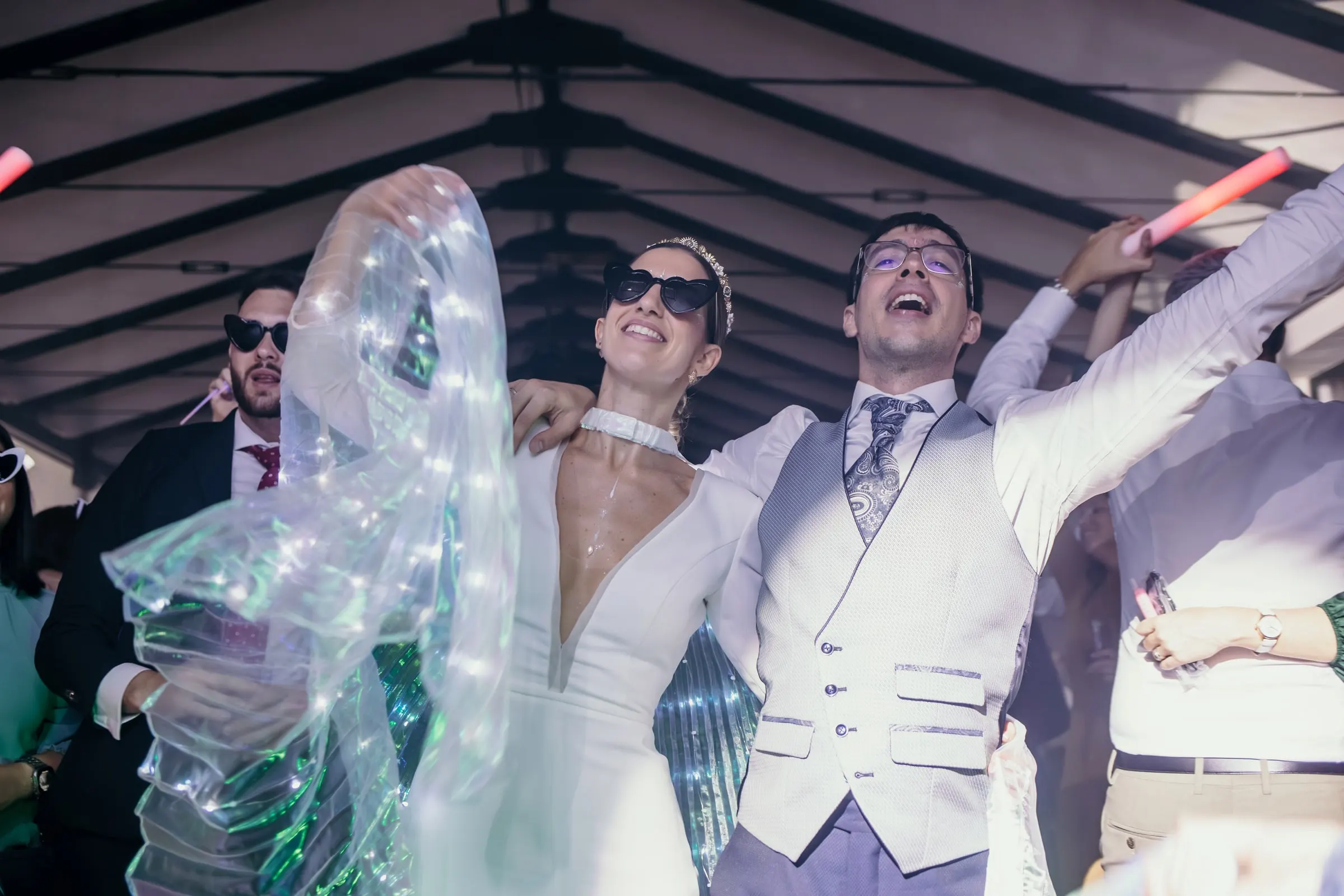 Bride with light-up wings and groom singing together in sunglasses at the party