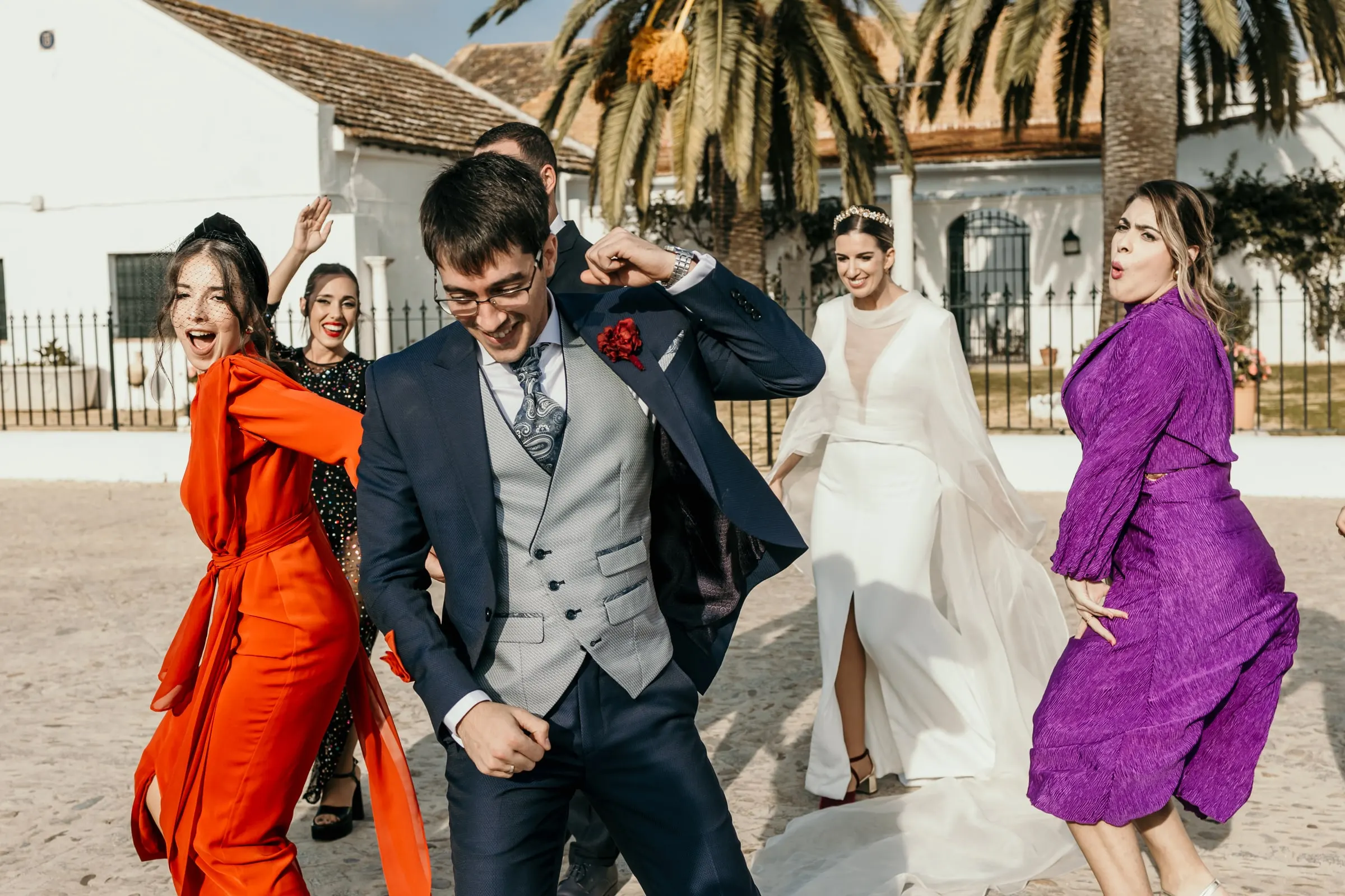 El novio baila con amigas de vestido naranja y morado en la explanada soleada del cortijo andaluz, con la novia detrás