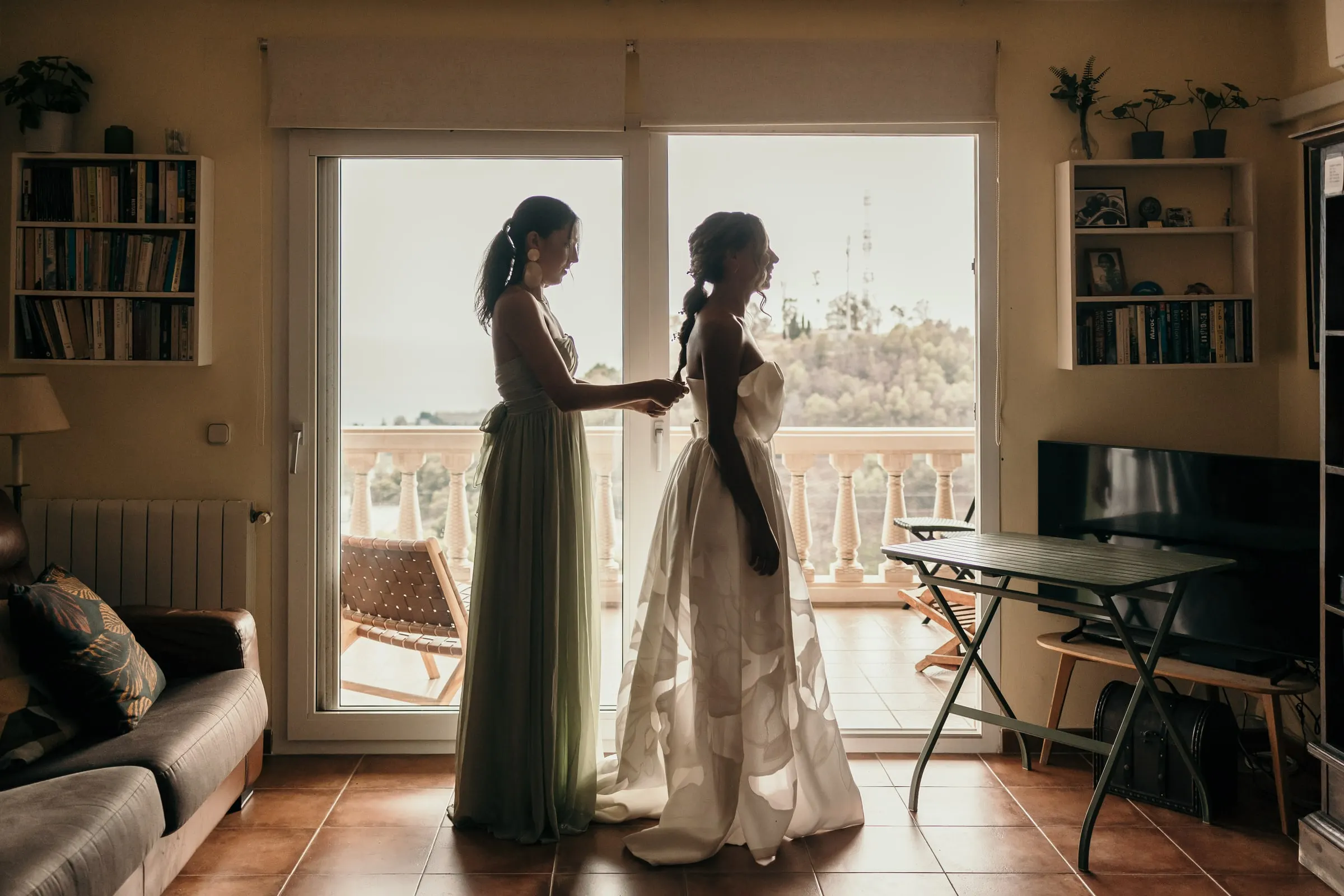 Una dama de verde salvia ayuda a la novia con el vestido junto a la puerta del balcón de una villa