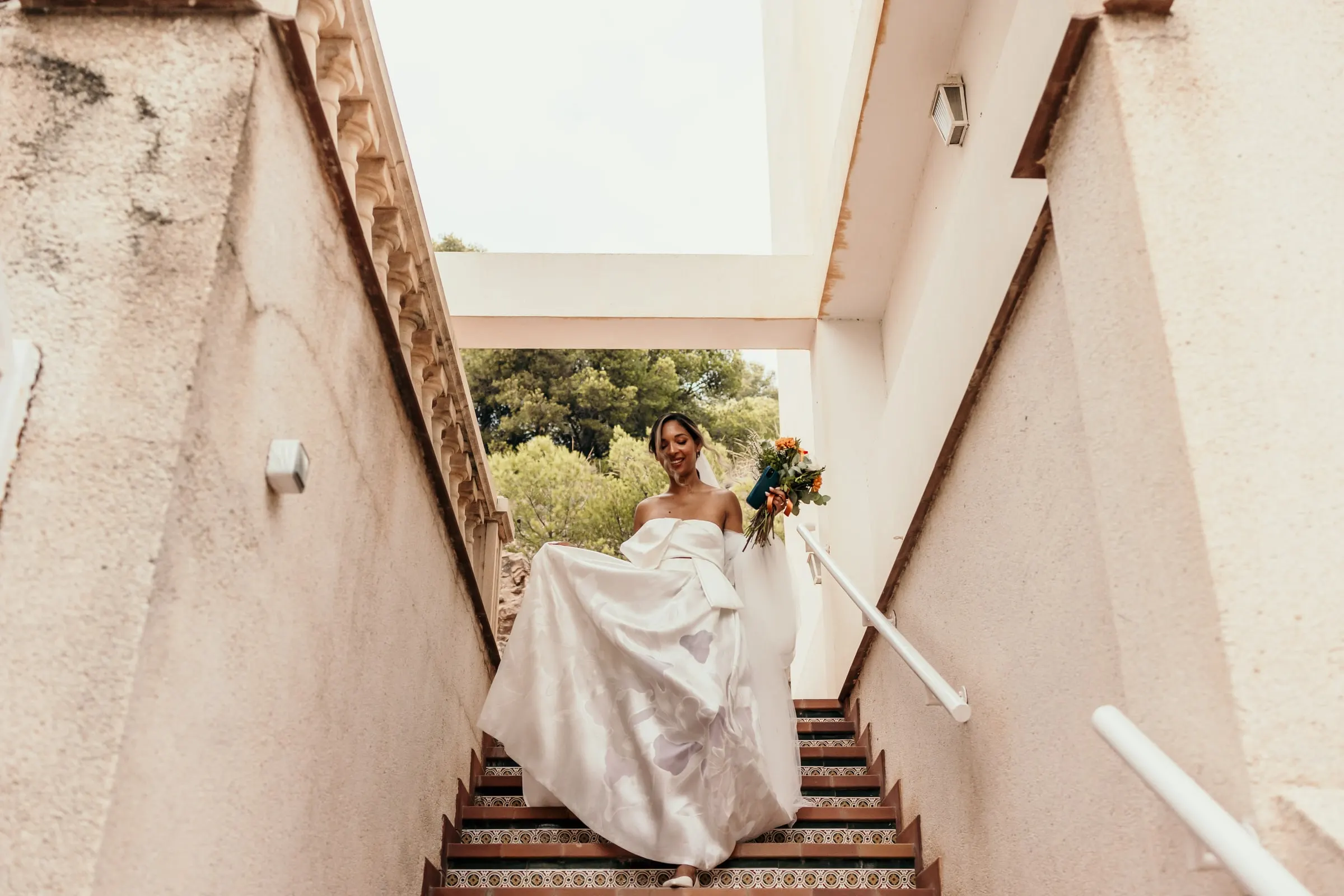 La novia se recoge la falda al bajar una escalera exterior entre paredes blancas
