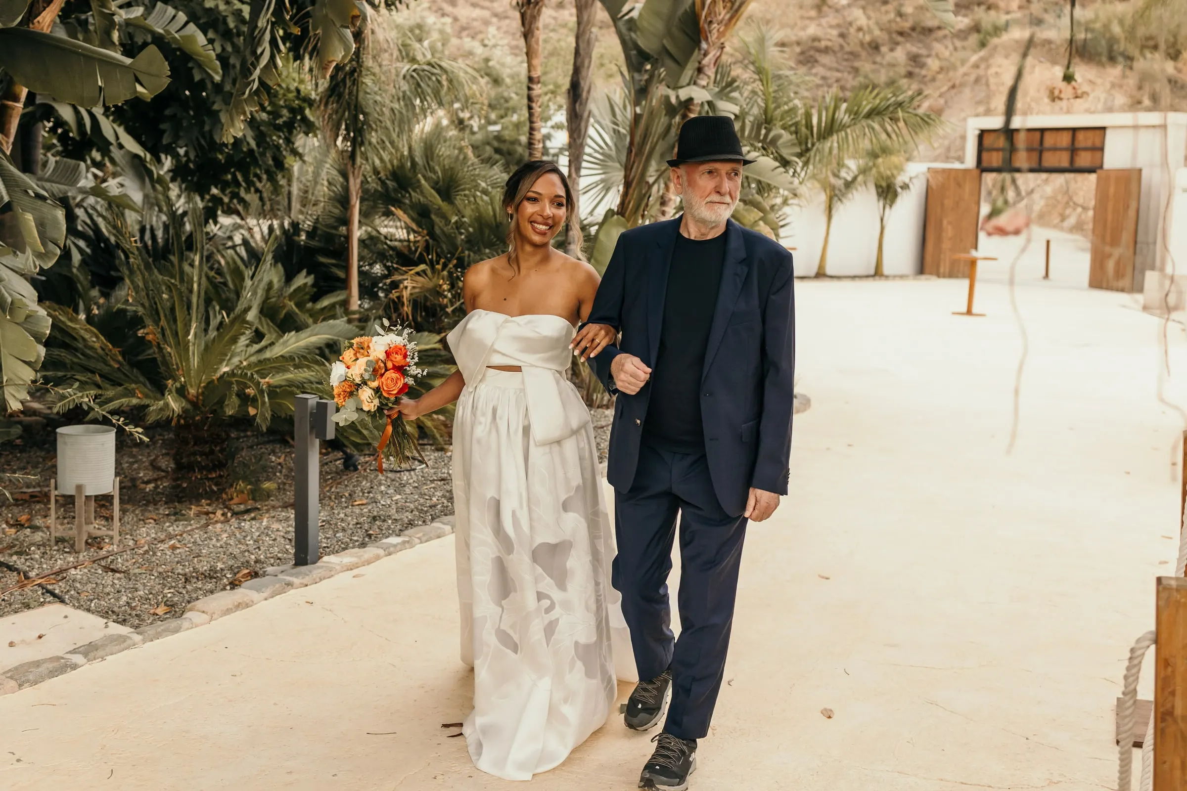 La novia llega del brazo del hombre del sombrero negro por un jardín tropical de palmeras