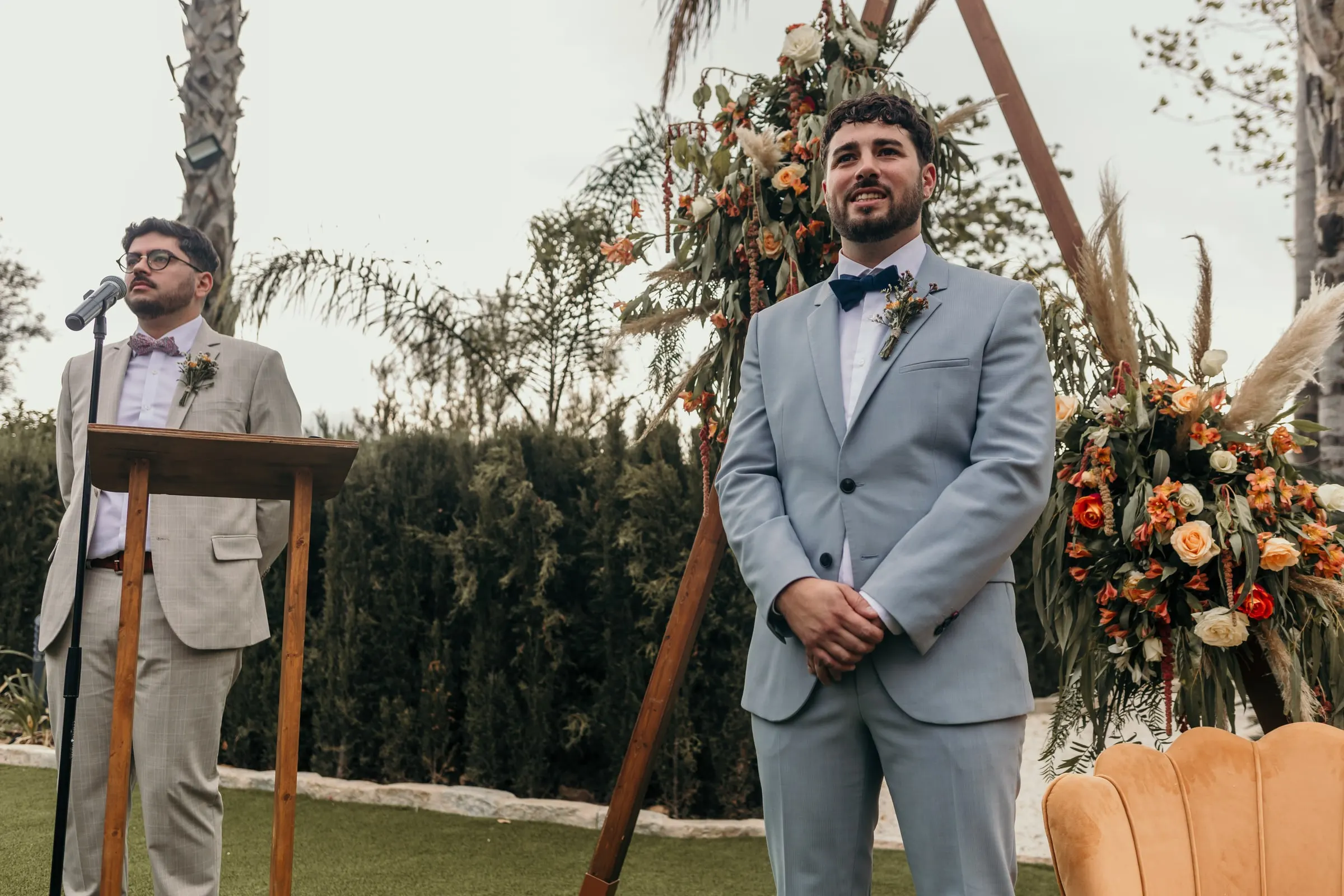 El novio, de traje azul claro y pajarita marino, espera junto al arco floral de la ceremonia al aire libre