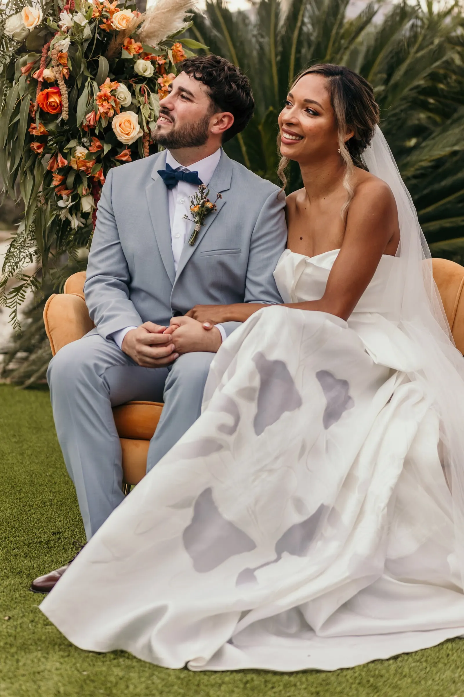 Los novios ríen en sillones de terciopelo junto al arreglo de flores naranjas