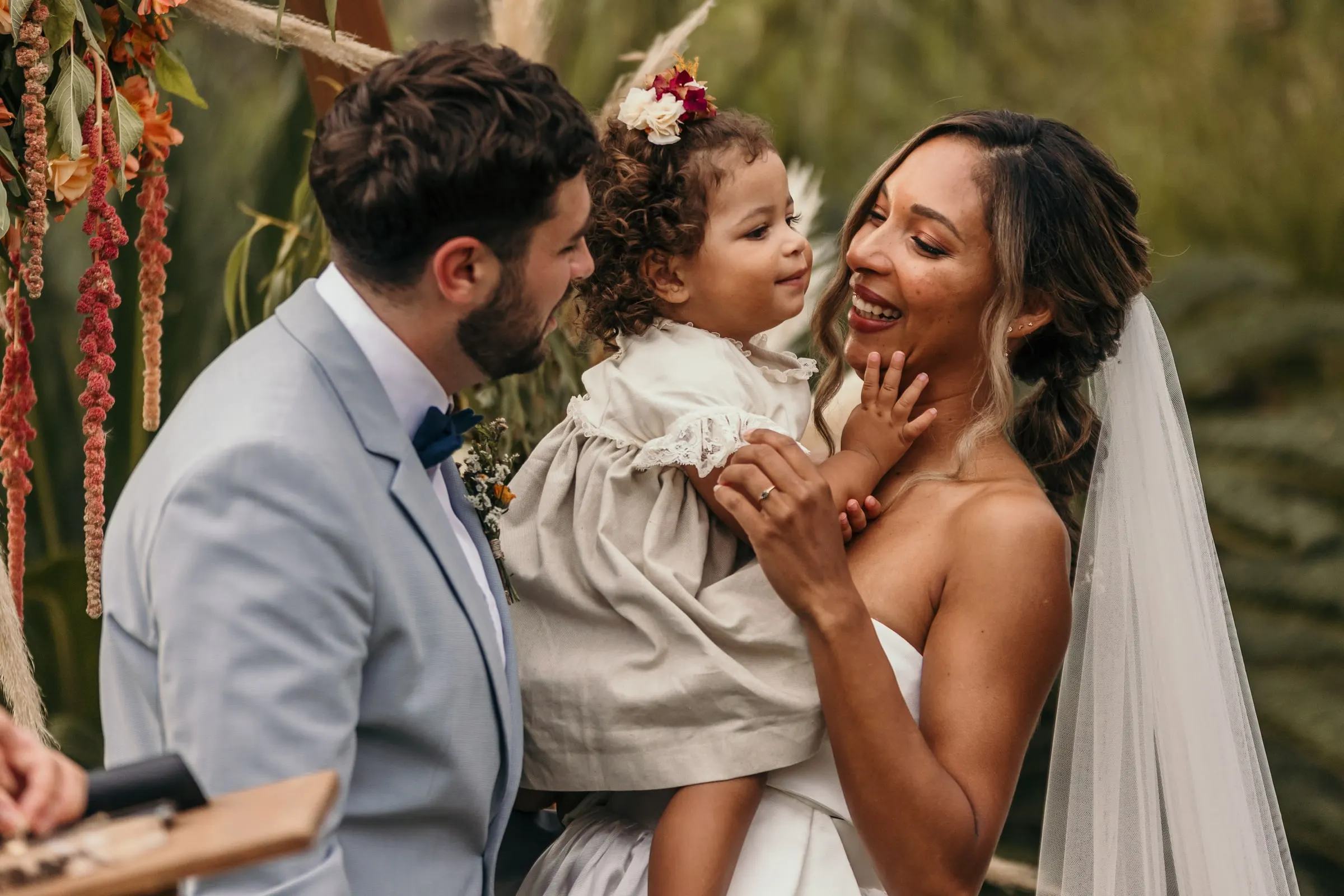 Los novios ríen con la niña de la corona de flores junto al arco floral