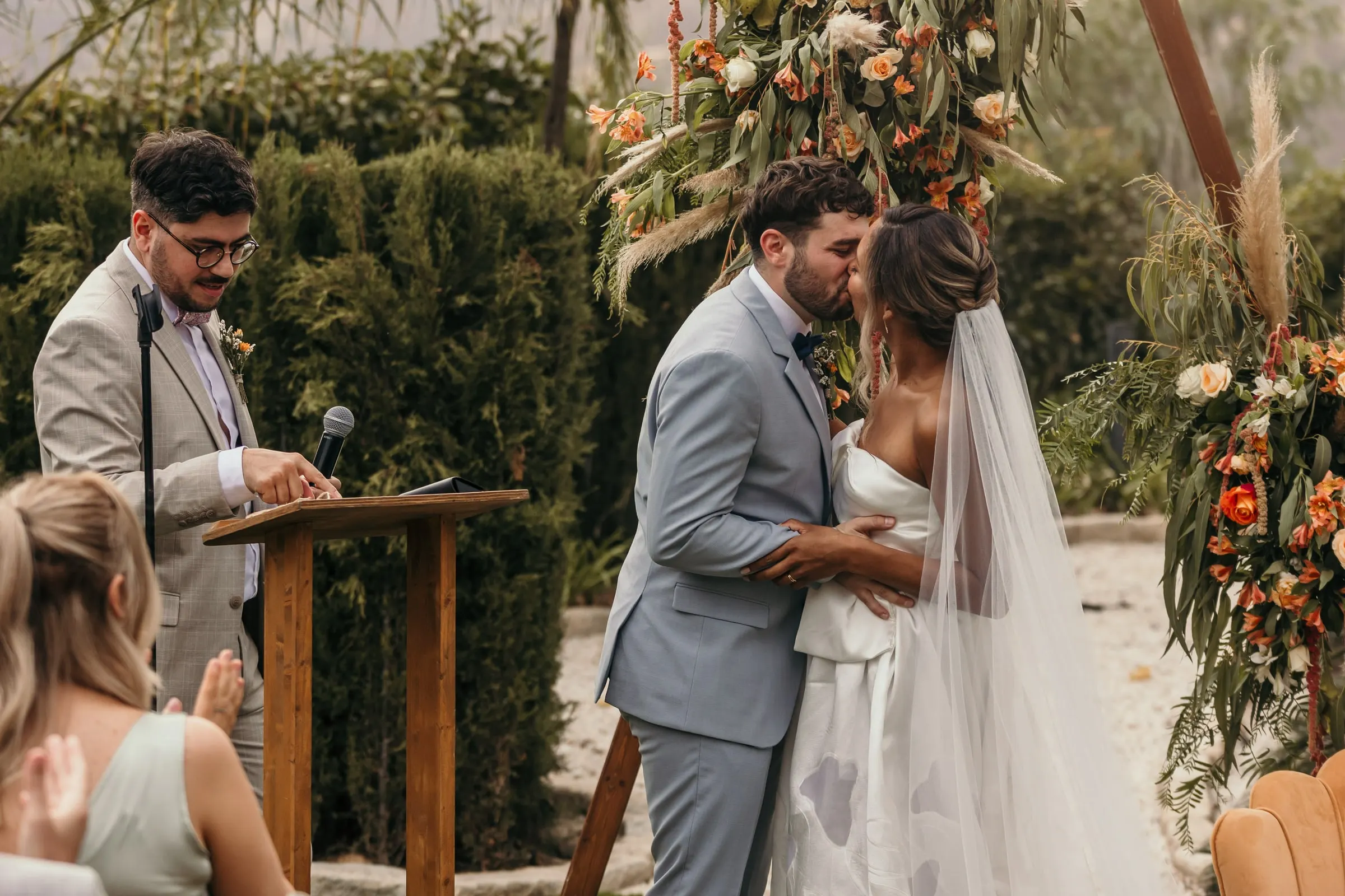 Primer beso en la ceremonia al aire libre junto al atril de madera y las flores naranjas