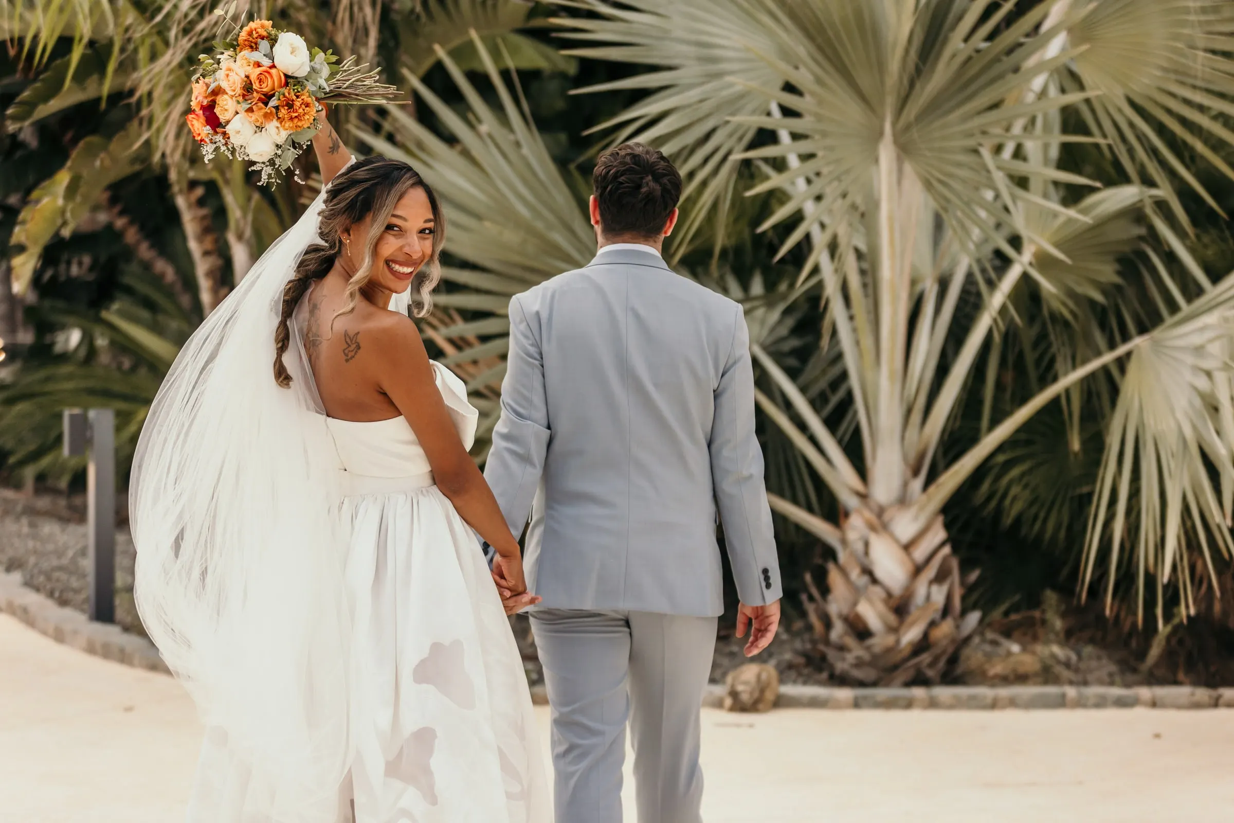 La novia mira atrás de la mano del novio entre palmitos