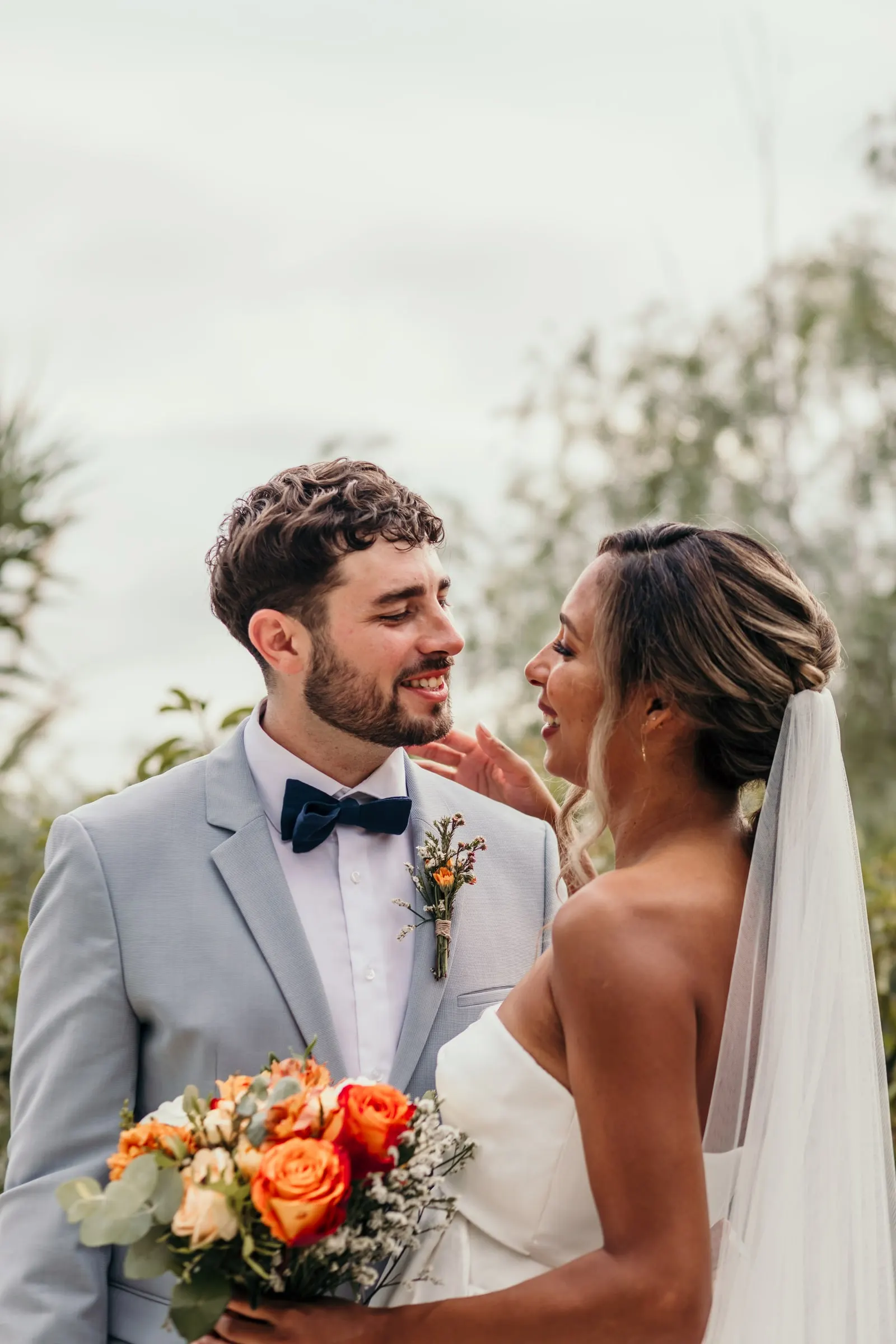 La novia toca la cara del novio mientras ríen, con las rosas naranjas en la mano