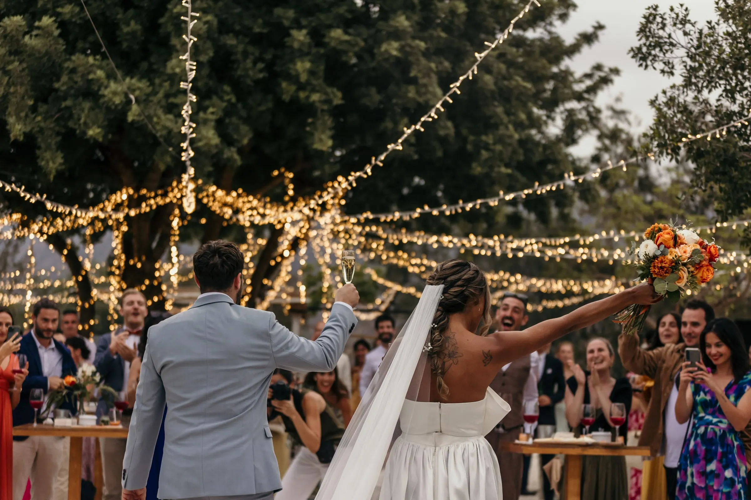 La novia alza el ramo ante los invitados bajo guirnaldas de luces