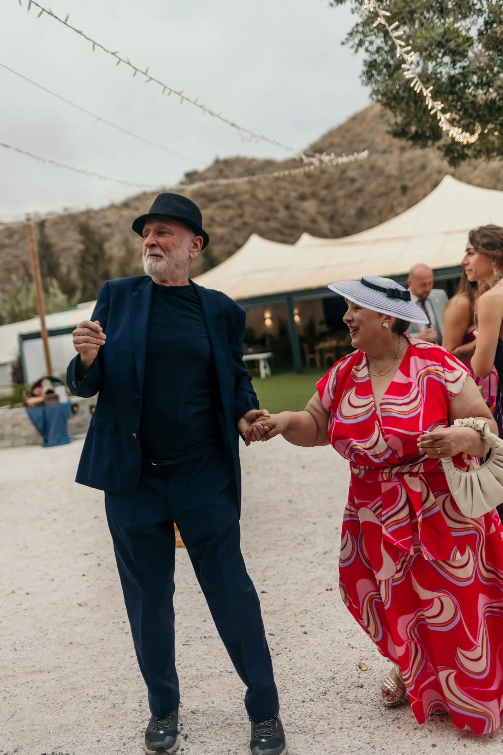 El hombre del sombrero negro baila con una invitada de vestido rojo y canotier ante una carpa blanca y un cerro seco