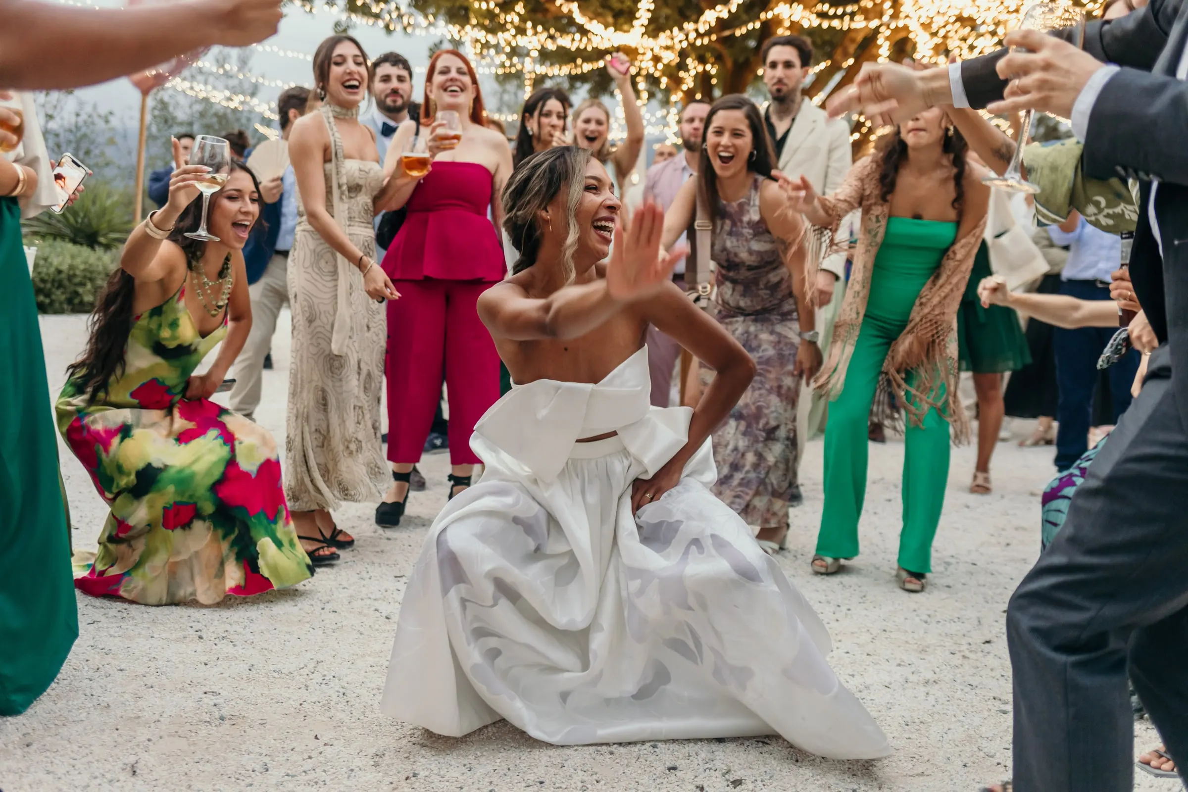La novia se agacha a bailar entre invitados de colores sobre la grava