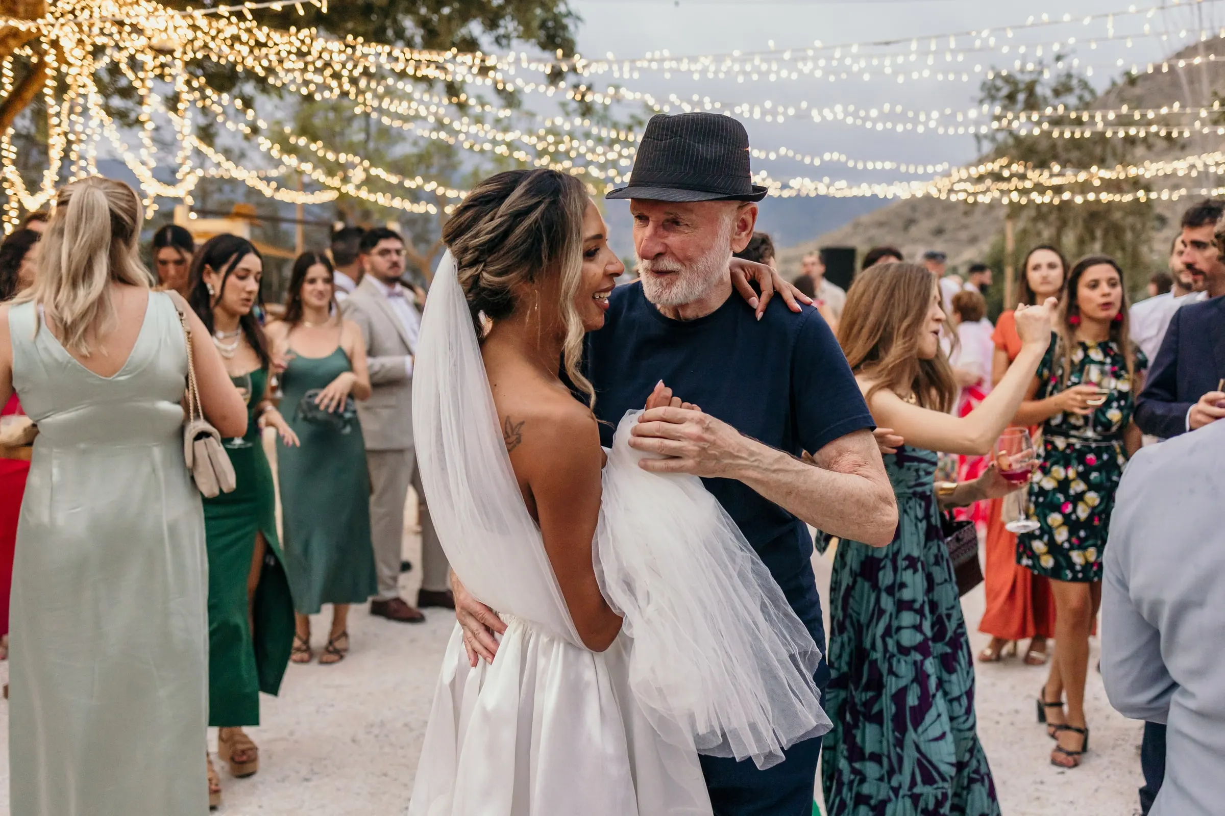 La novia ríe bailando con el hombre del sombrero negro bajo las luces