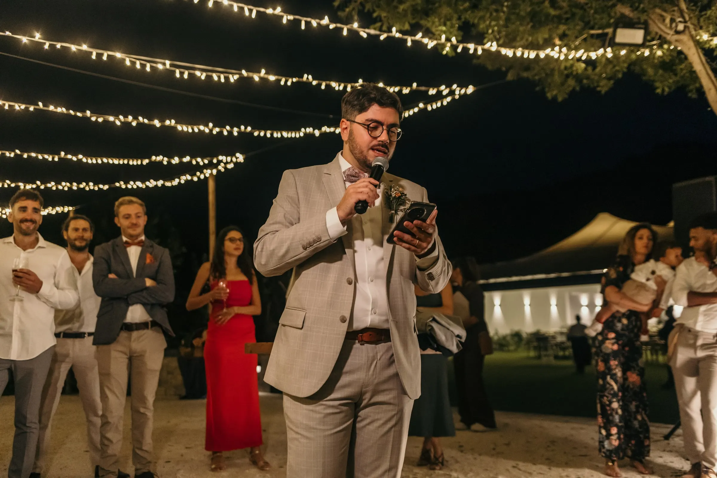 Un amigo del novio, de traje beige, habla al micrófono en la cena al aire libre