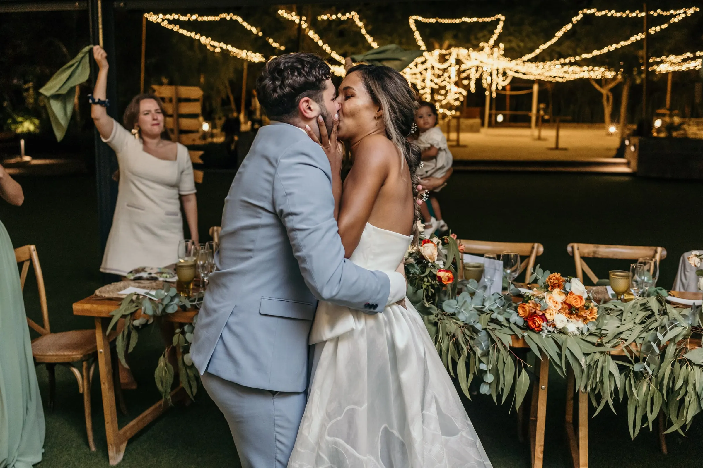 Los novios se besan junto a la mesa larga decorada con verde y flores naranjas