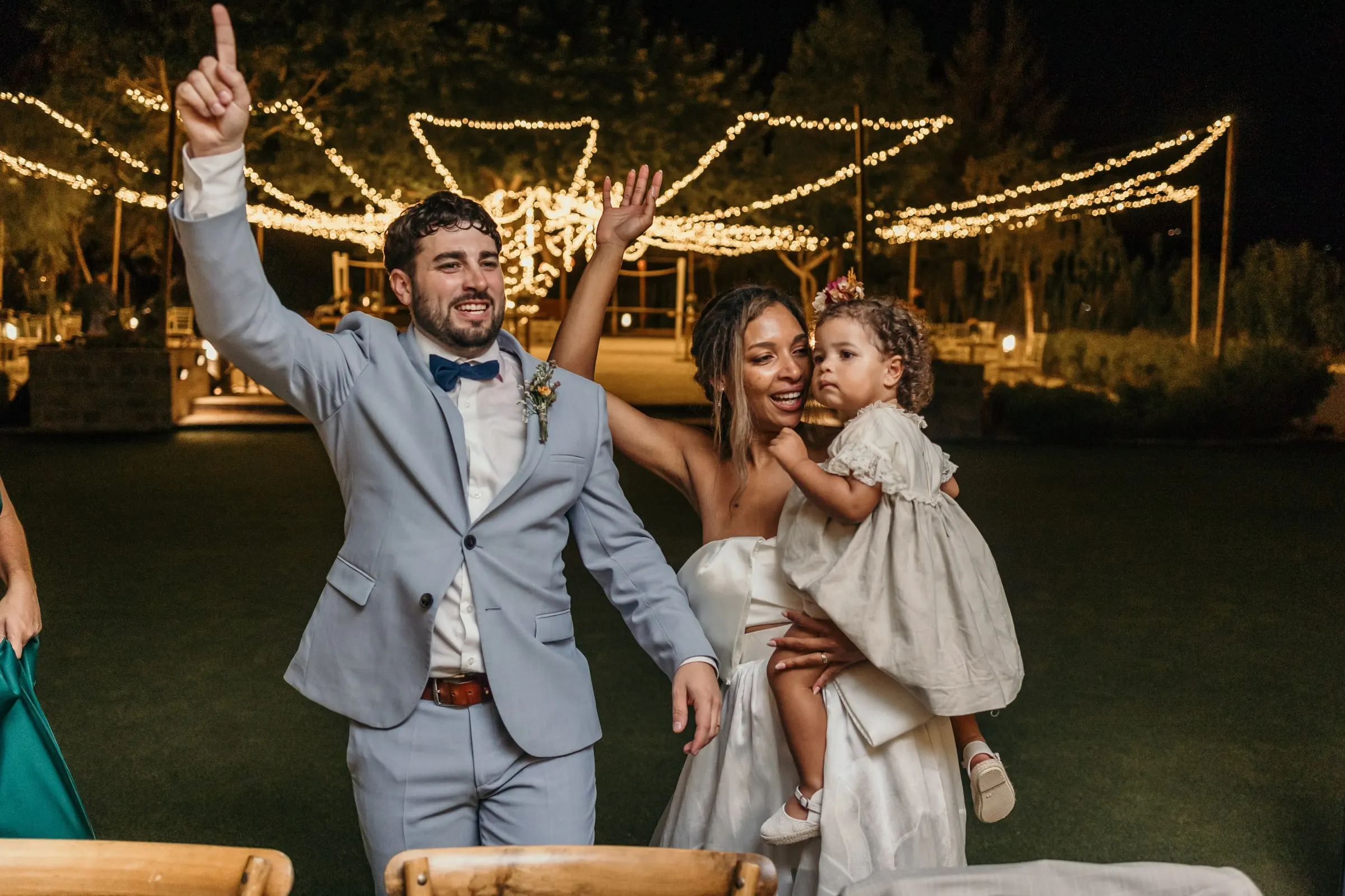 El novio levanta el brazo mientras la novia sostiene a la niña, con las guirnaldas detrás