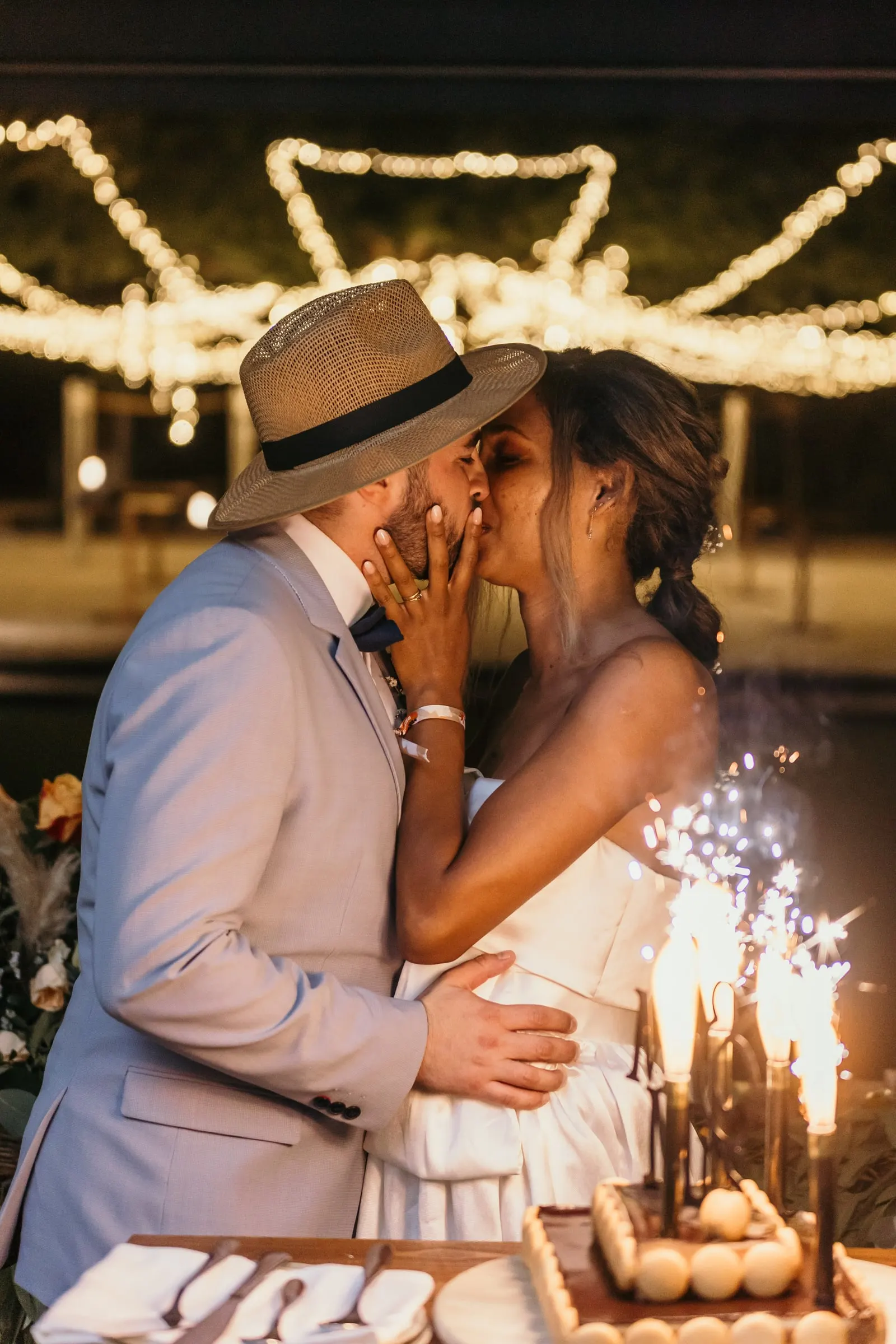 El novio con sombrero de paja besa a la novia junto a las bengalas de la tarta