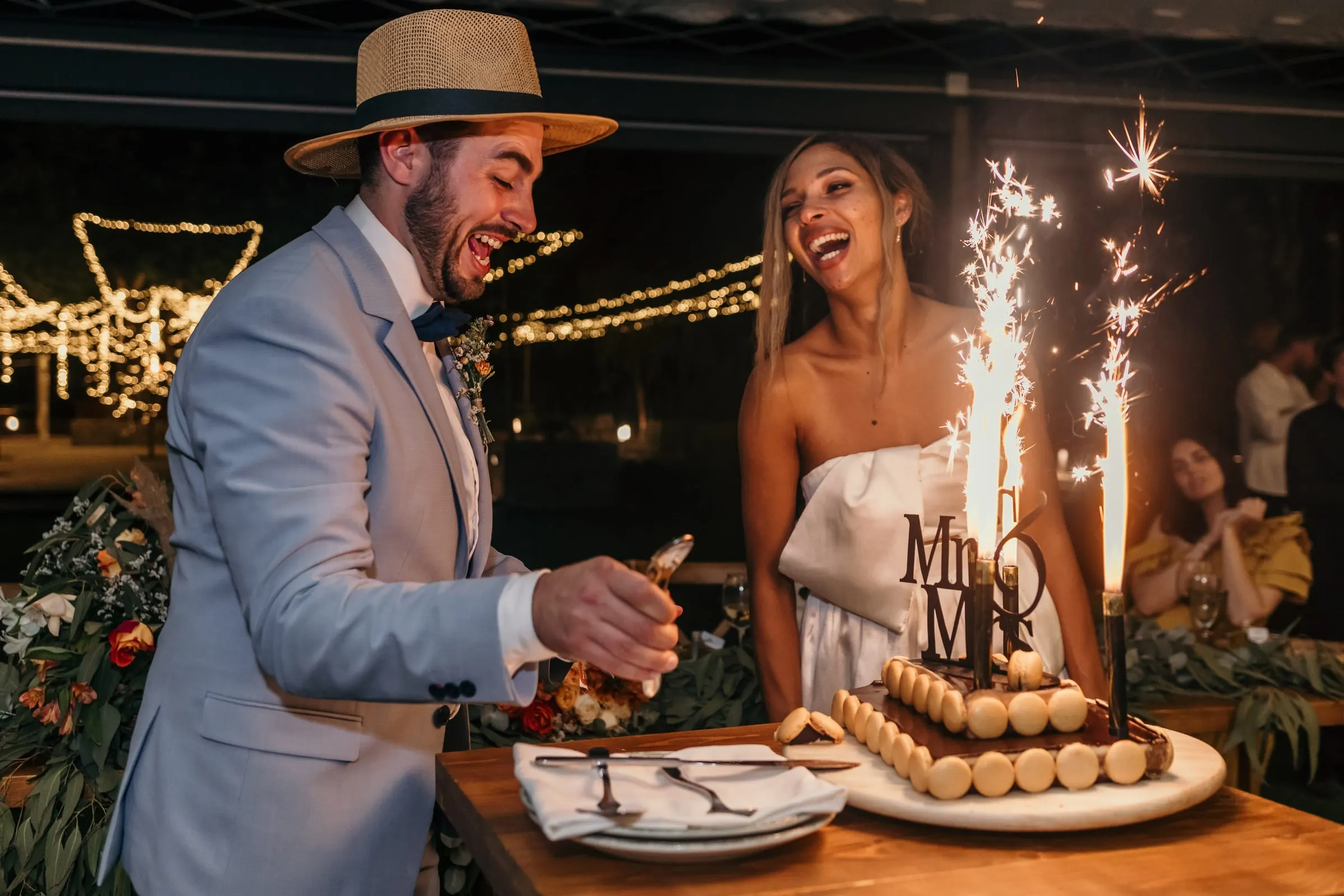 Los novios ríen al cortar una tarta con bengalas y figura de Mr & Mrs