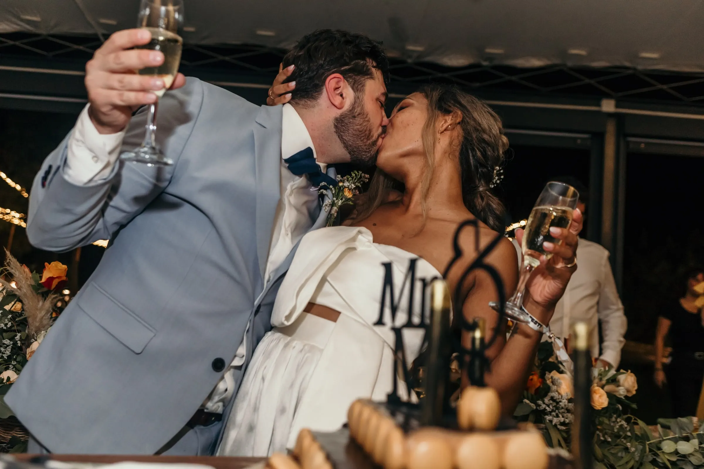 Los novios se besan con las copas de cava en alto junto a la mesa de la tarta